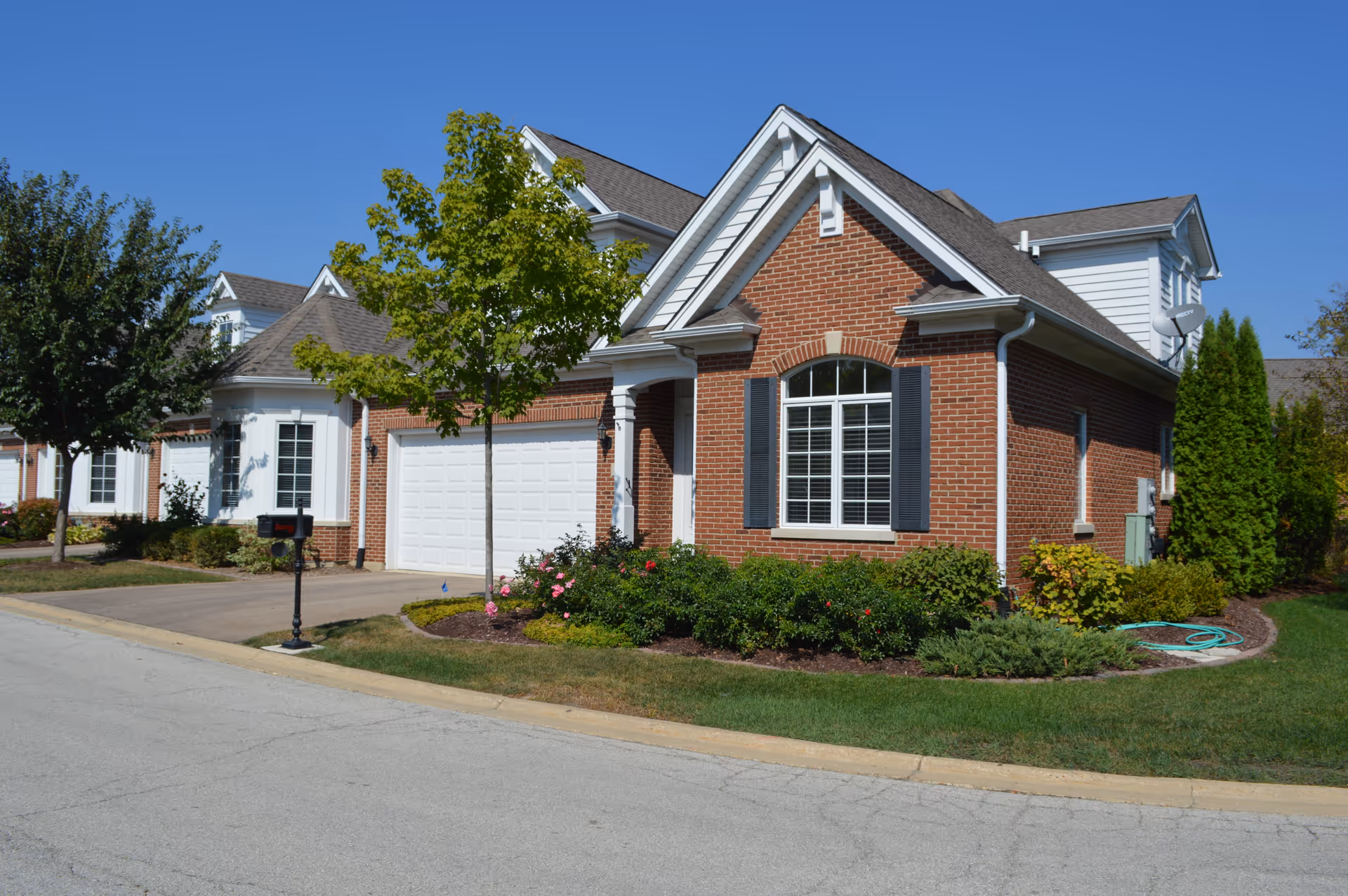 Brick senior living facility with driveway, attached garage, and manicured landscaping under a blue sky.
