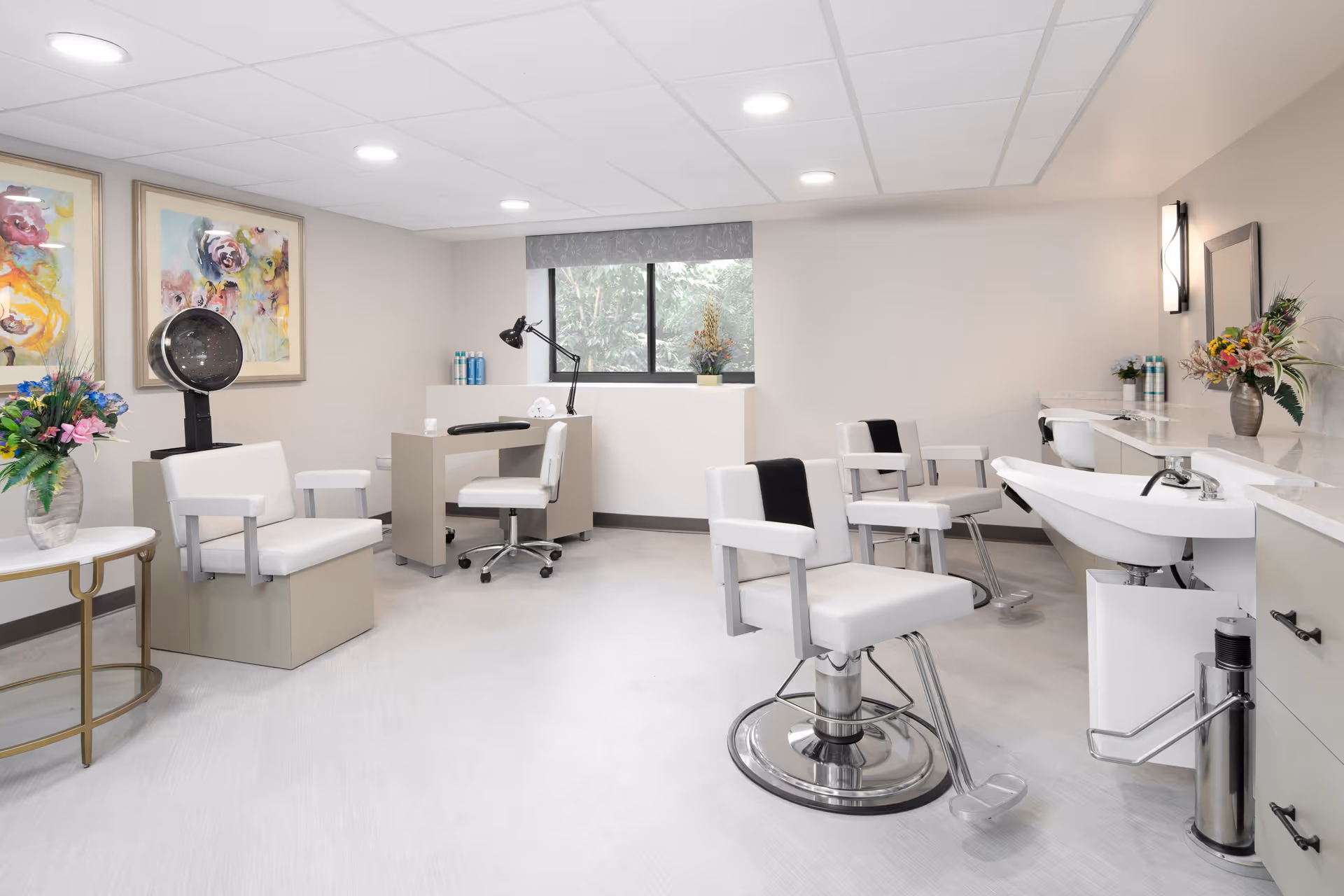 A bright and clean salon area in a senior living facility with white salon chairs, a hair dryer, a manicure station, a large sink, and floral decorations. The room has light-colored walls, a window with a gray valance, and colorful abstract paintings on the wall.