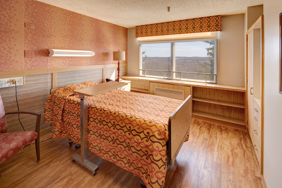 A patient bedroom in a senior care facility with a patterned bed, chair, and large window.