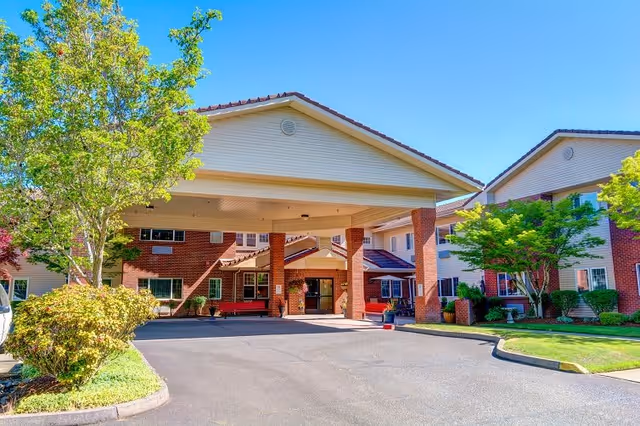 Front exterior view of The Lodge at Vista View building with a covered entrance, brick and siding facade, surrounded by green trees and shrubs under a clear blue sky.