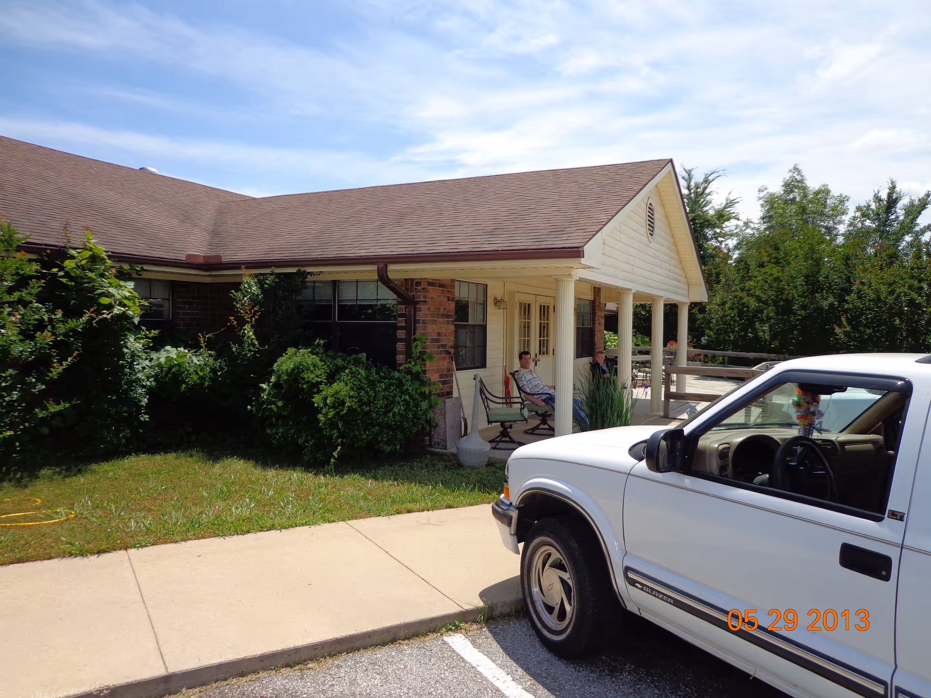 Front of a single-story brick-and-siding building with a covered porch where two people sit and a white pickup truck parked in front.