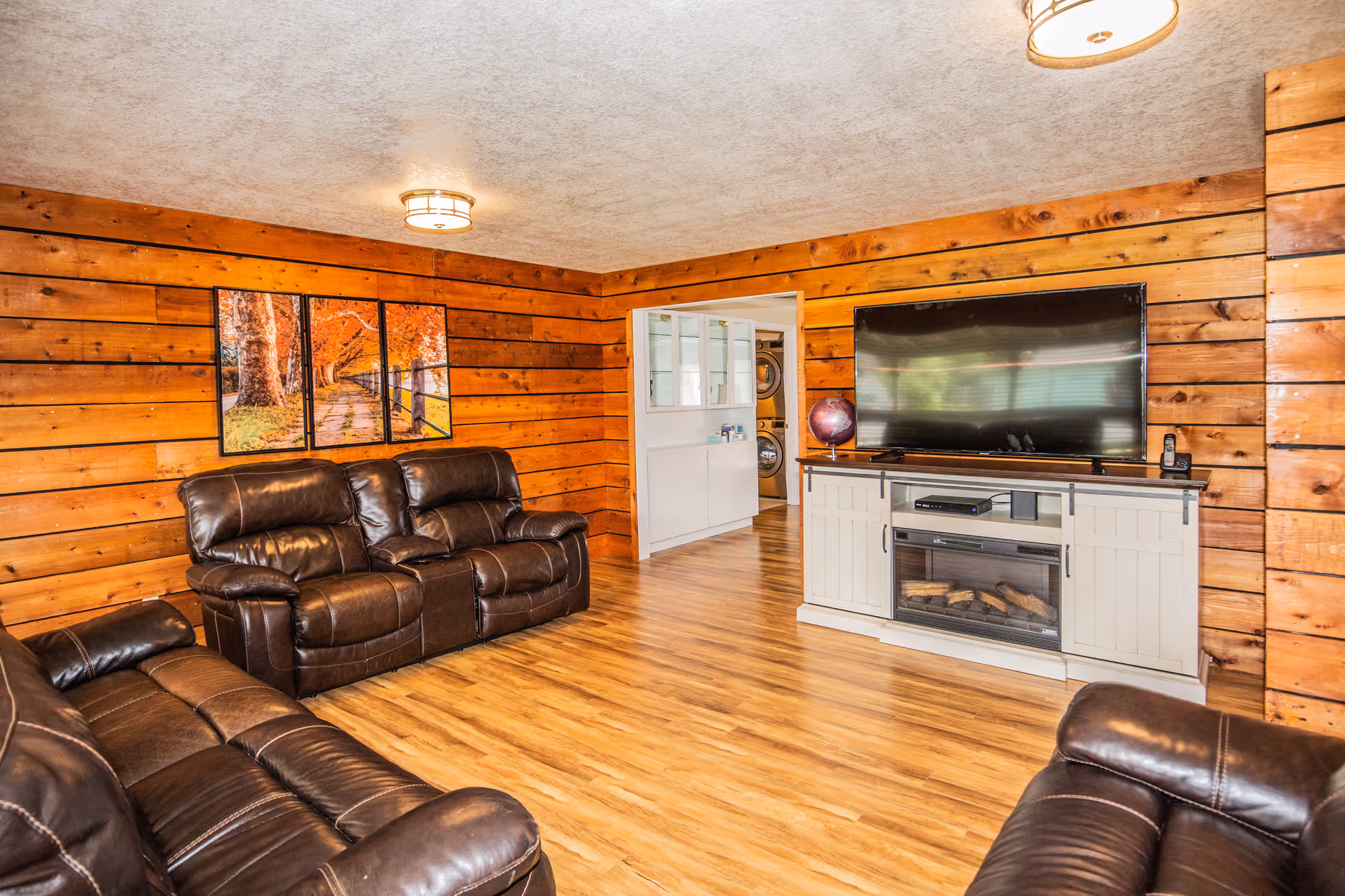 Cozy living room with wood-paneled walls, leather recliner sofas, a large flat-screen TV on a cabinet and wood-look flooring.