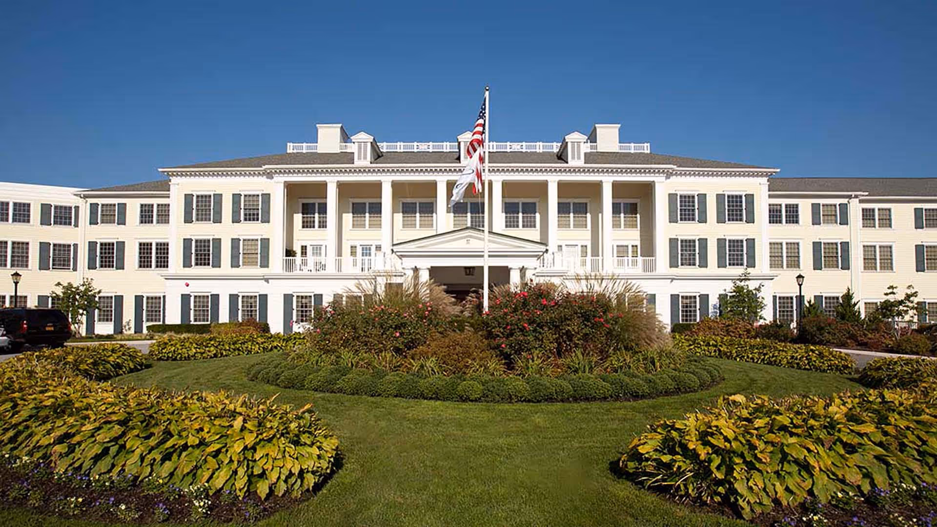 Large white three-story senior living building with columns, a landscaped circular driveway, and an American flag in front under a clear blue sky.