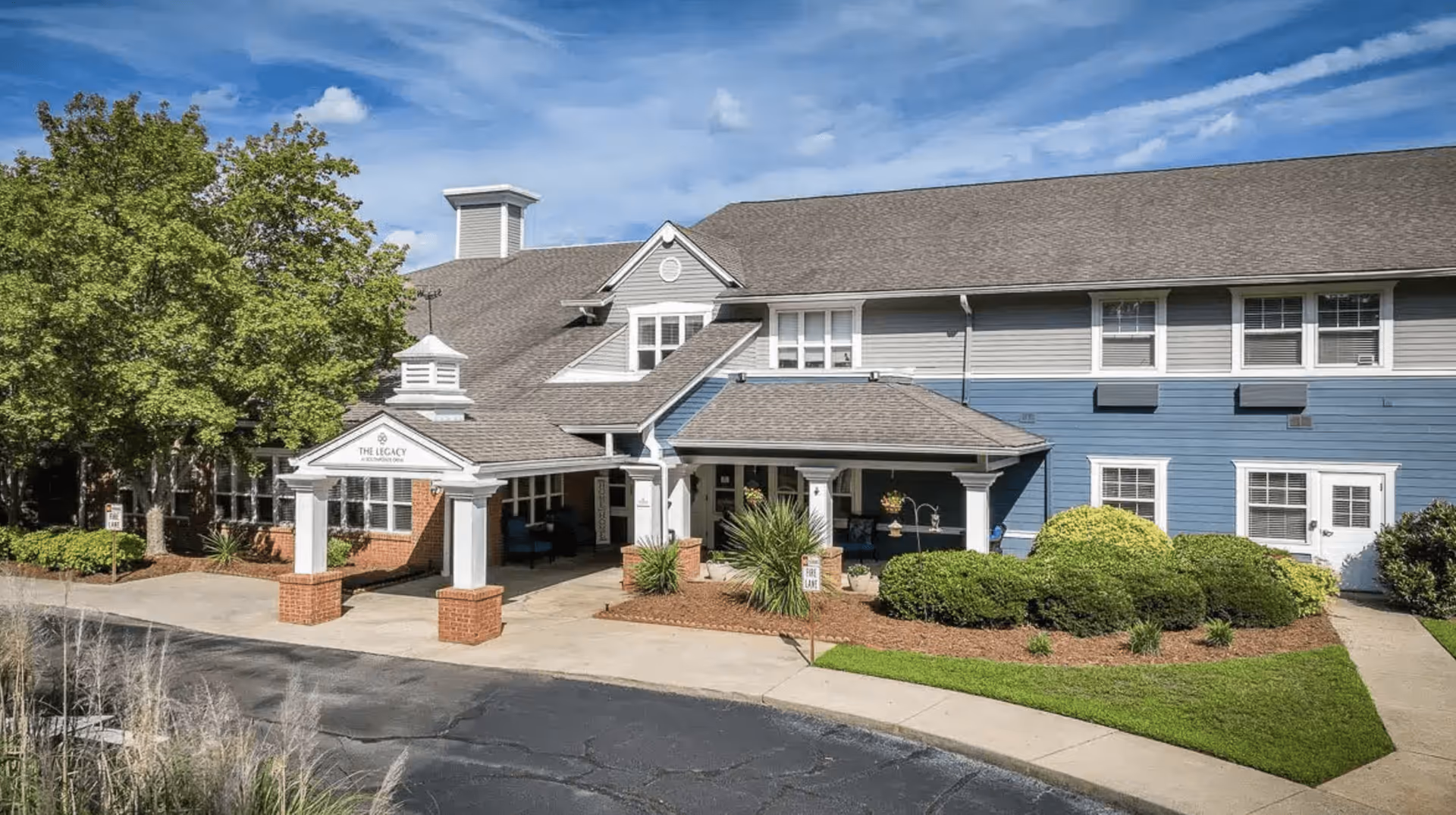 Exterior view of The Legacy at Southpointe Drive senior living facility showing a two-story building with blue and gray siding, a covered entrance with white columns and brick bases, surrounded by green bushes and trees under a partly cloudy sky.