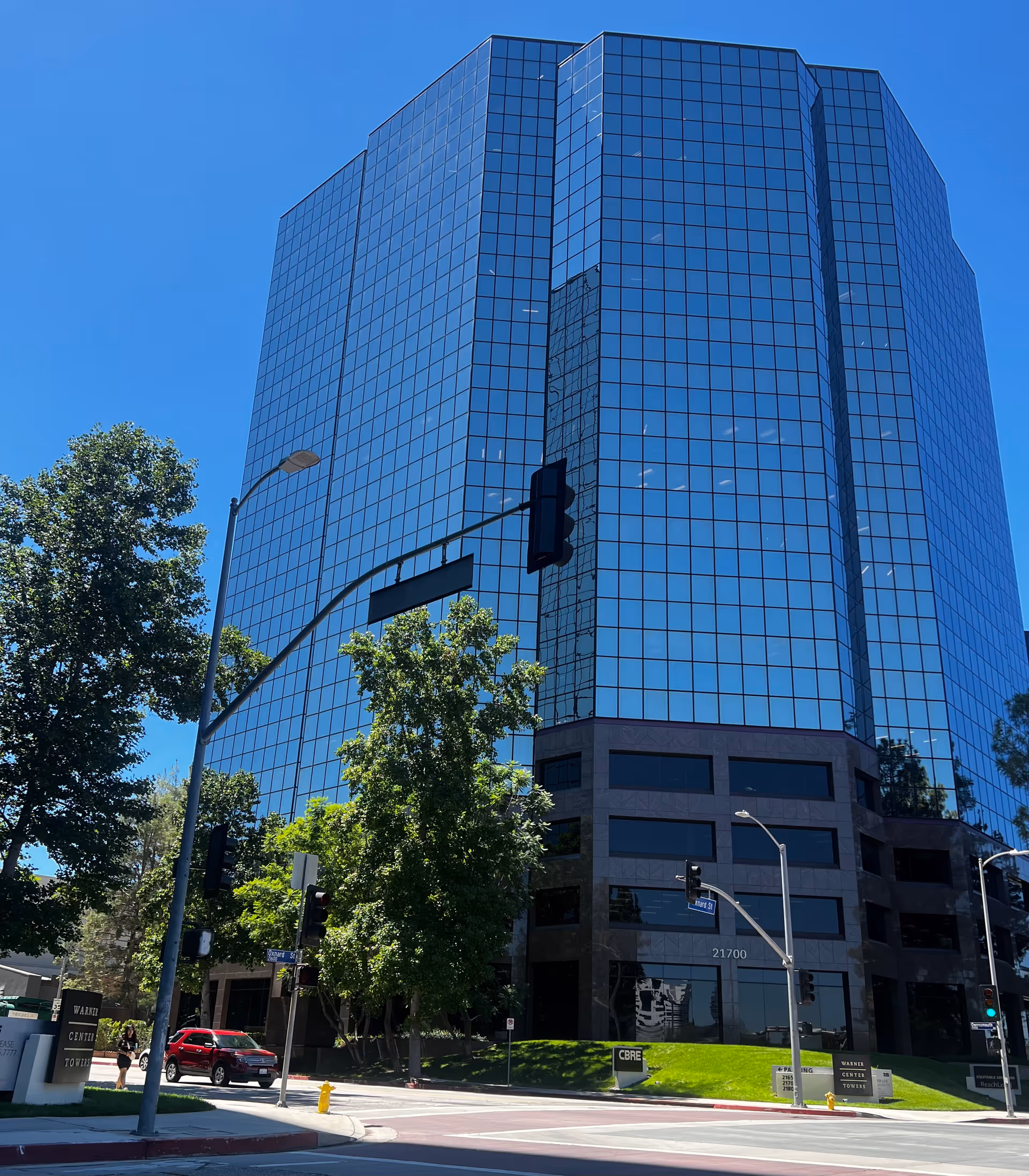 Tall, reflective glass office building on a sunny street with traffic signals and trees.