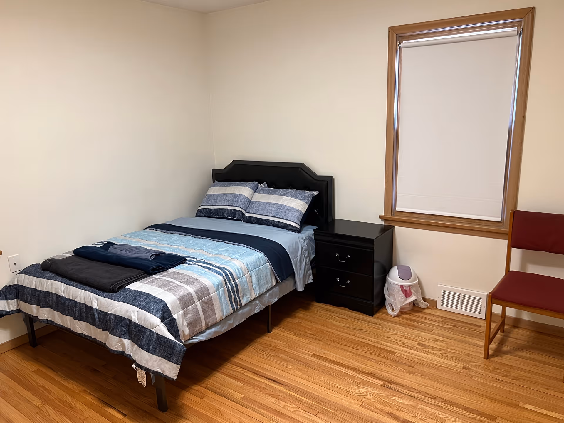 A small bedroom in a senior facility featuring a bed, nightstand, window, and chair.