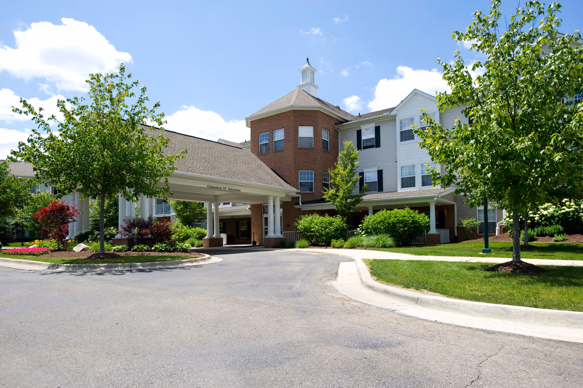 Front entrance of a multi-story senior living building with a covered porte-cochère, landscaped grounds, and trees under a blue sky.