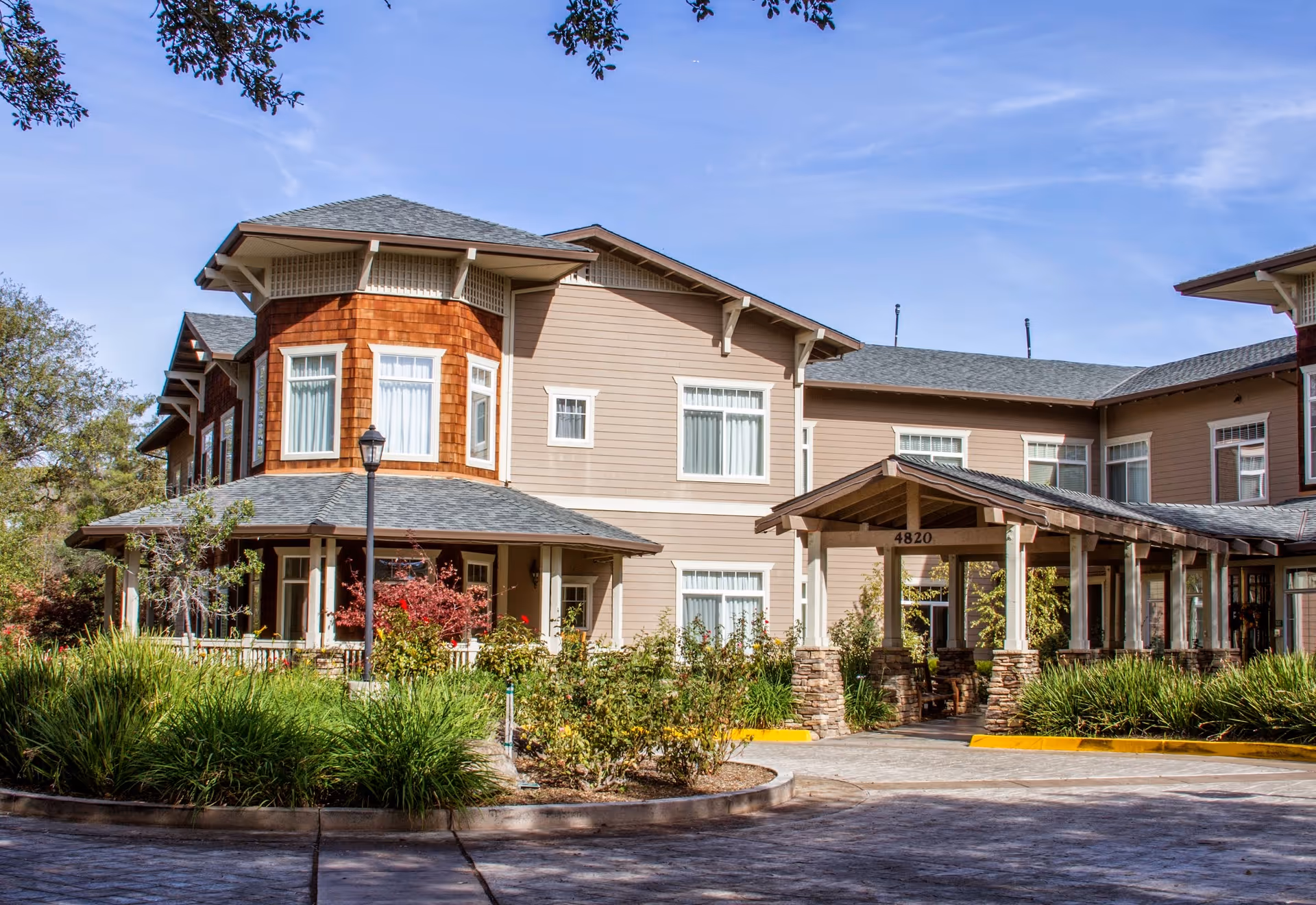 Front exterior of a two-story senior living building with a covered entrance, landscaped grounds, and the address '4820' visible above the porte-cochere.