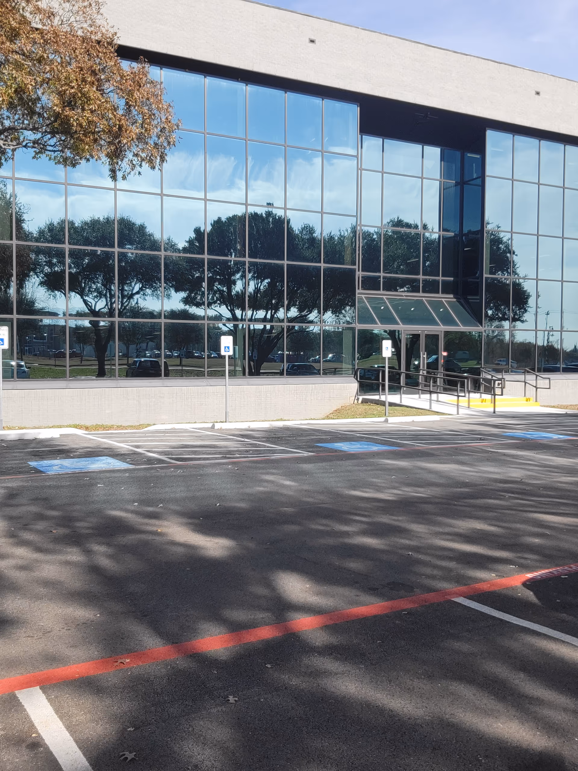 Modern glass-front facility exterior with ramp, parking, and reflected trees in windows.