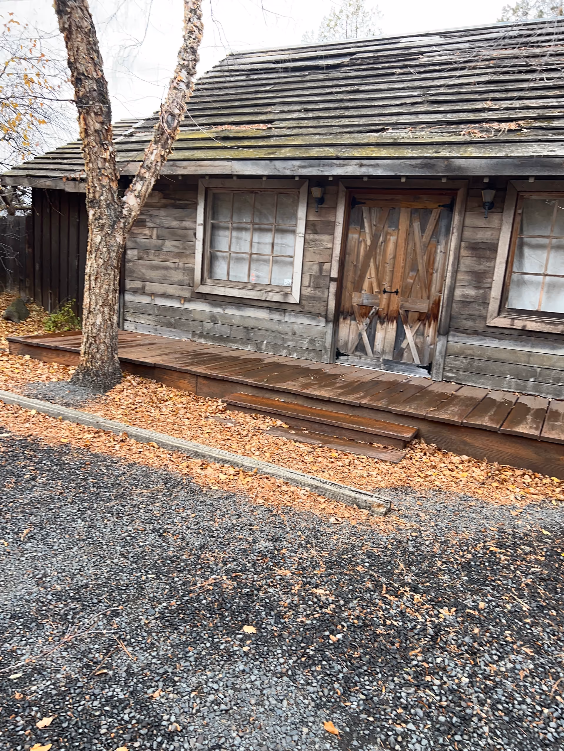 Weathered wooden building with a double door, two windows, and fallen leaves surrounding.