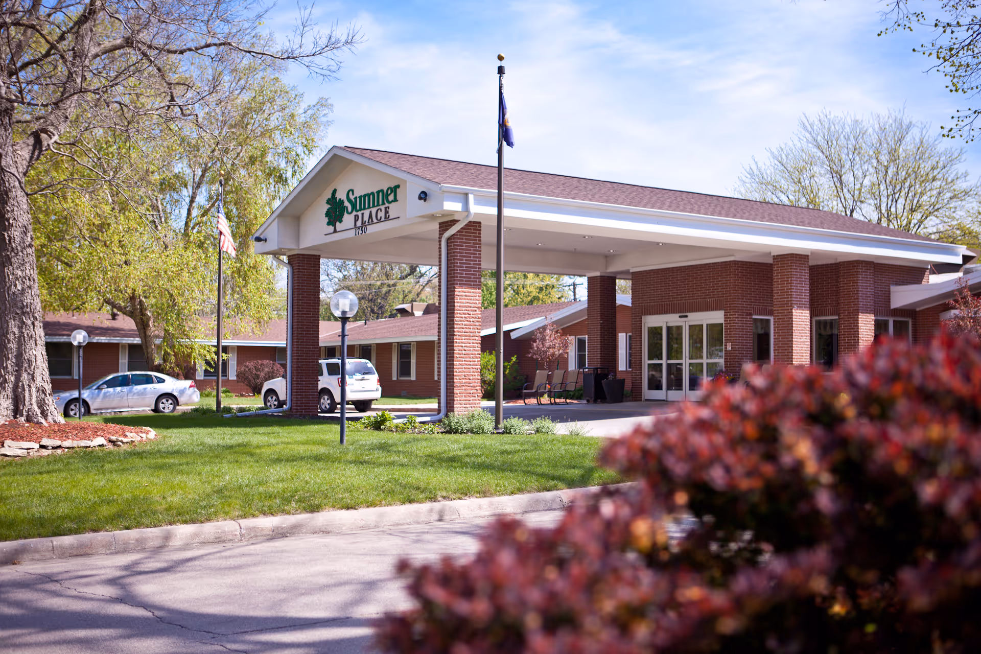 Front entrance of Sumner Place skilled nursing facility, a one-story brick building with a covered drive-through entrance, flagpoles, lawn, and parked cars.