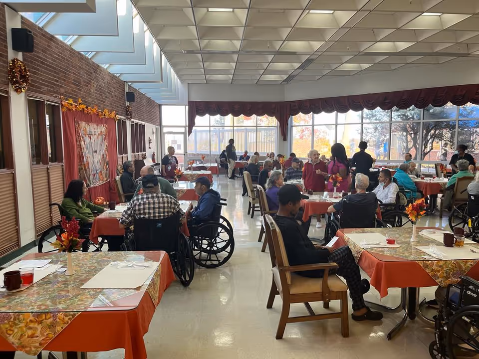 Residents in wheelchairs and seniors dine at tables in a bright, busy dining room.