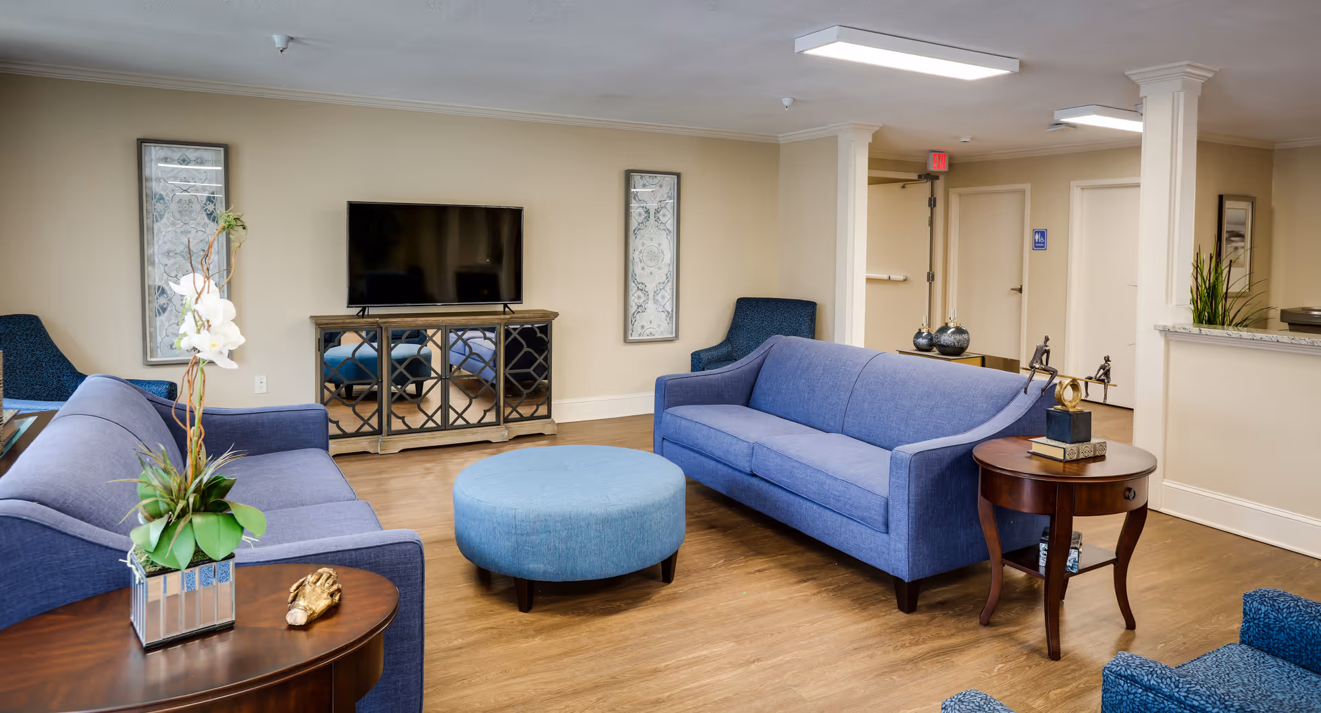 A cozy living room area with two blue sofas, a round blue ottoman, and a wooden side table with decorative items. A flat-screen TV is mounted on a mirrored cabinet against the wall, flanked by two framed artworks. The room has wooden flooring and neutral-colored walls with a doorway and hallway visible in the background.