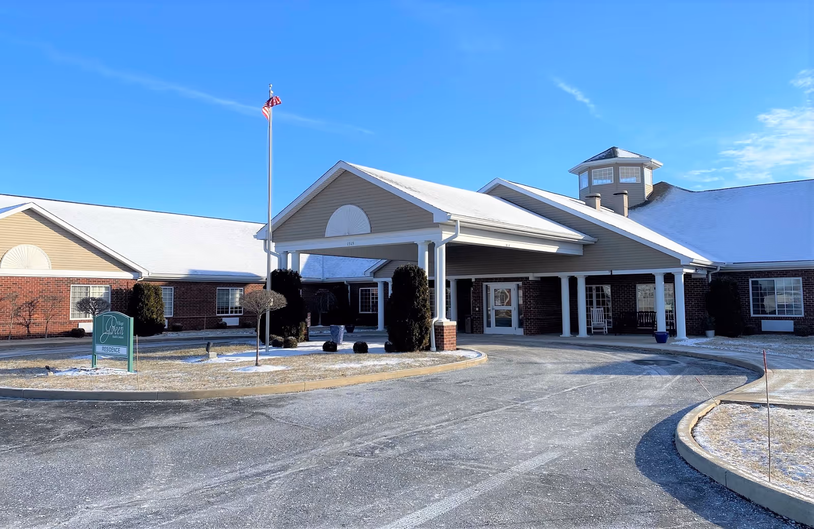 Village Green Health Campus exterior with entrance canopy, flagpole, and blue sky.