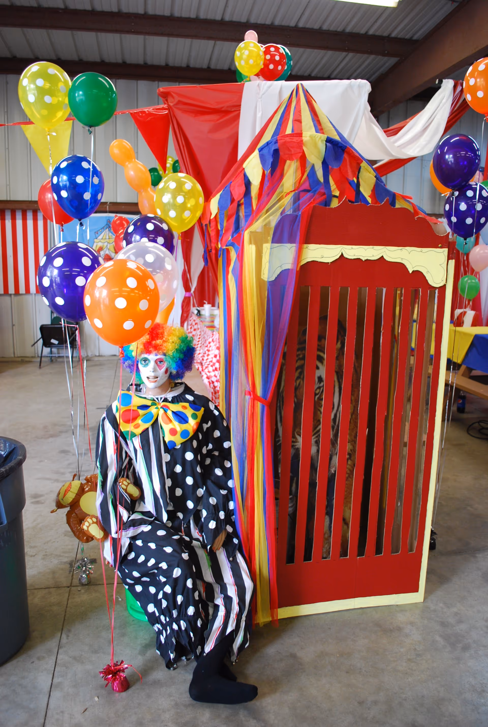 Clown figure seated beside a colorful carnival booth inside a decorated facility.