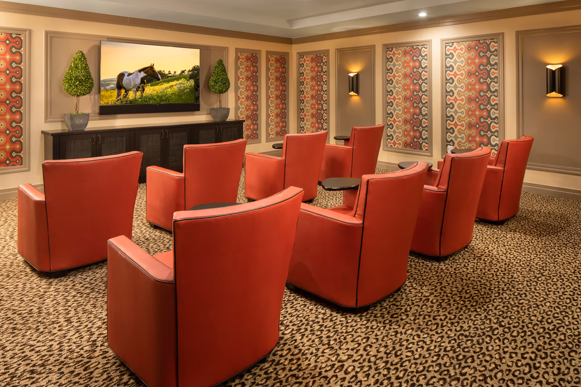 A cozy media room with eight red armchairs arranged in two rows facing a large flat-screen TV mounted on the wall. The TV displays an image of a horse in a field. The room has patterned carpet, decorative wall panels with colorful geometric designs, two potted topiary plants on a dark wooden cabinet beneath the TV, and wall sconces providing warm lighting.