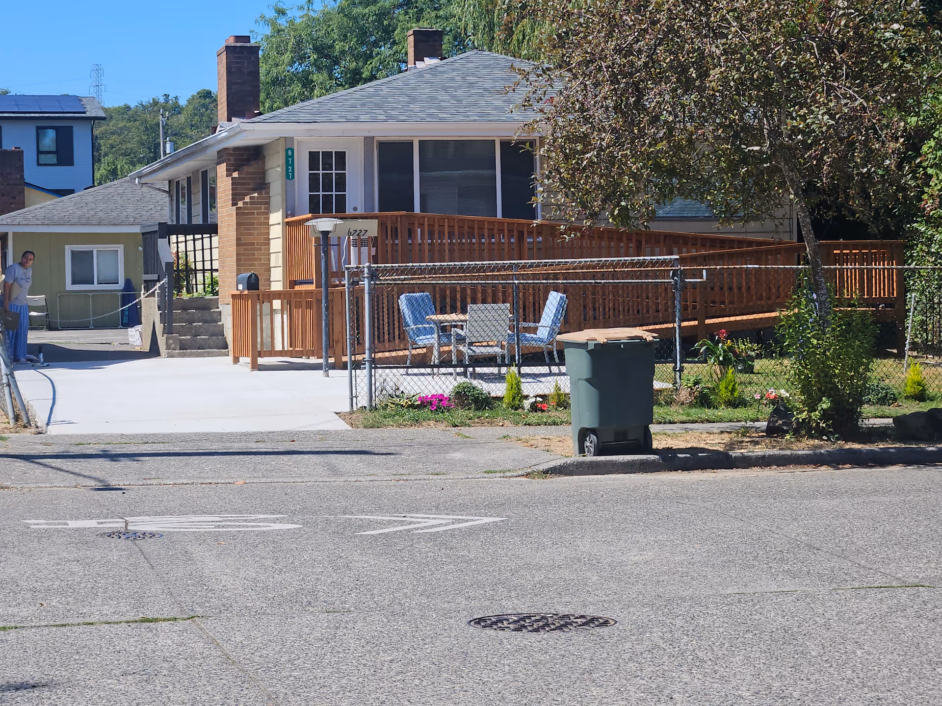 Single-story house with wooden deck, outdoor seating, fence, trash bin, and a person nearby.