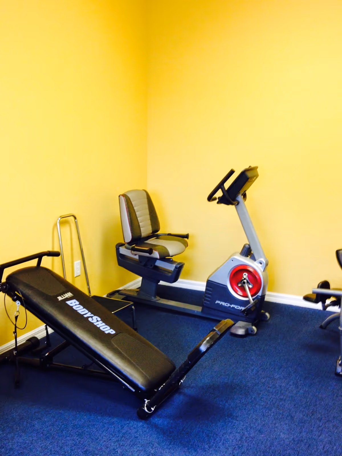 Yellow-walled interior with gym equipment: treadmill, recumbent bike, and workout bench.