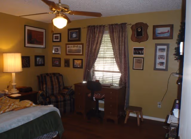 Cozy bedroom with bed, desk, chair, window with curtains, and wall photo gallery.