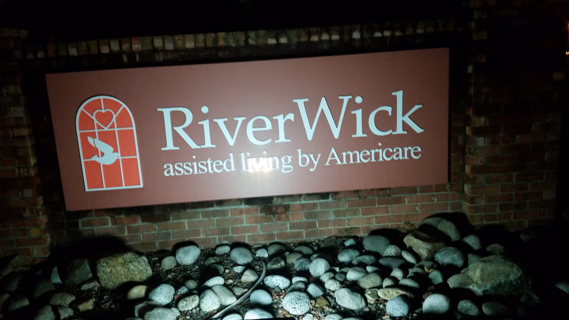 Exterior brick wall with RiverWick assisted living sign and decorative rocks at base.