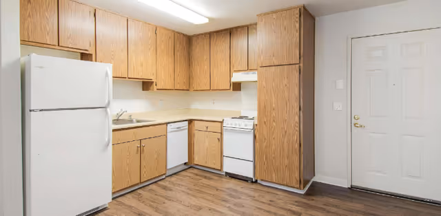 Compact kitchen with wooden cabinets, white appliances, sink, stove, and dishwasher.
