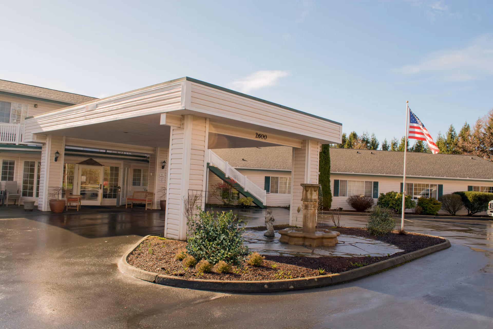 White senior living facility entrance with porte-cochere, flagpole, fountain, and landscaped circle drive.