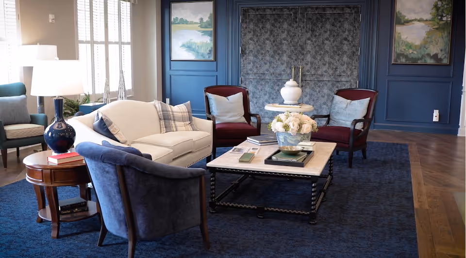A comfortable seating area in a senior living facility featuring sofas and armchairs around a coffee table with a floral centerpiece against blue paneled walls.