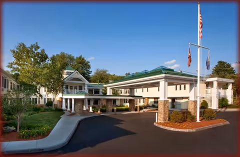 Exterior view of Hanover Hill Health Care Center showing a large building with a covered entrance, multiple flags on flagpoles, landscaped greenery, and a clear blue sky.