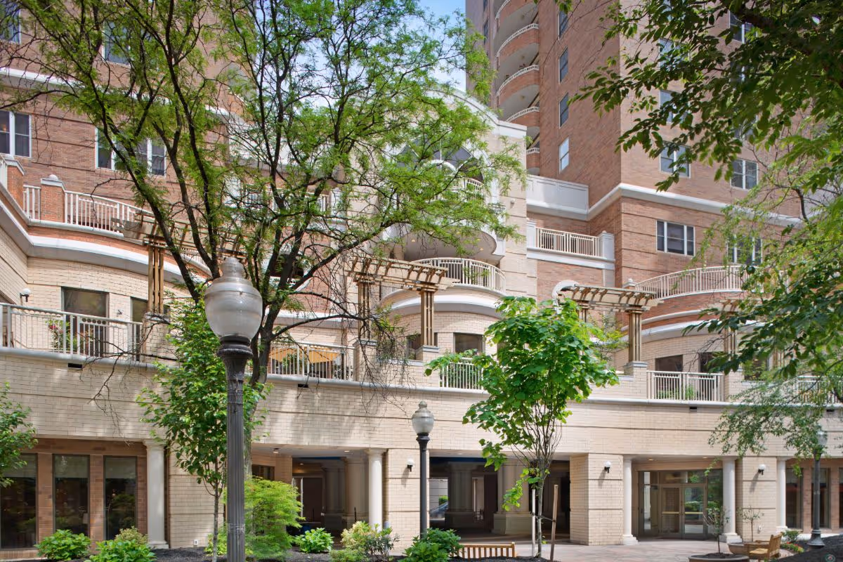 Front courtyard and entrance of a multi-story brick residential building with balconies, trees, and lampposts.