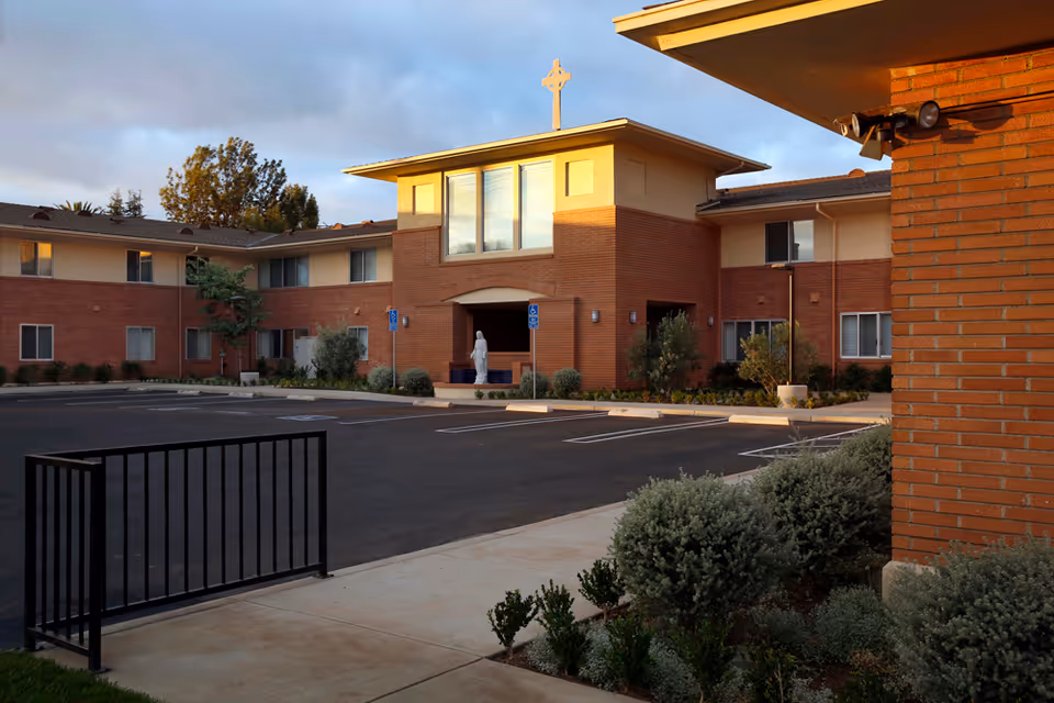 Brick two-story building front with a cross above the entrance, a statue by the doorway, an empty parking lot, and landscaped shrubs.