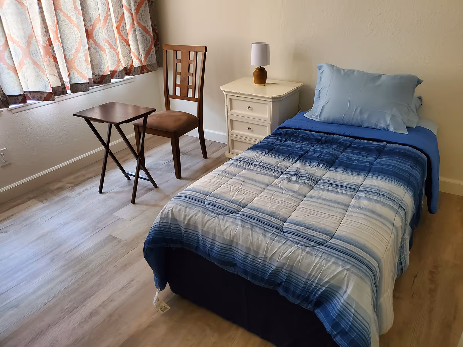 Bedroom in a senior living facility with a bed, nightstand, chair, and window.