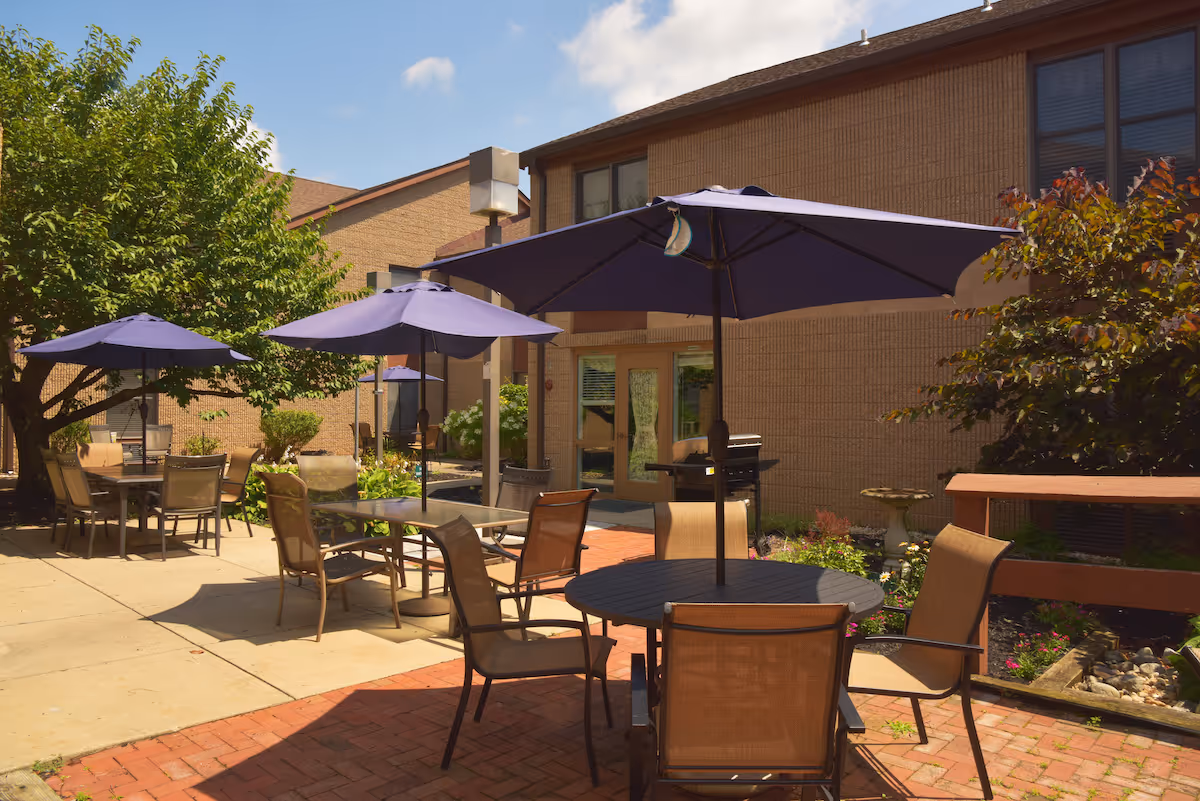 Outdoor patio area with several tables and chairs under large purple umbrellas, surrounded by trees and plants, adjacent to a brick building under a partly cloudy sky.