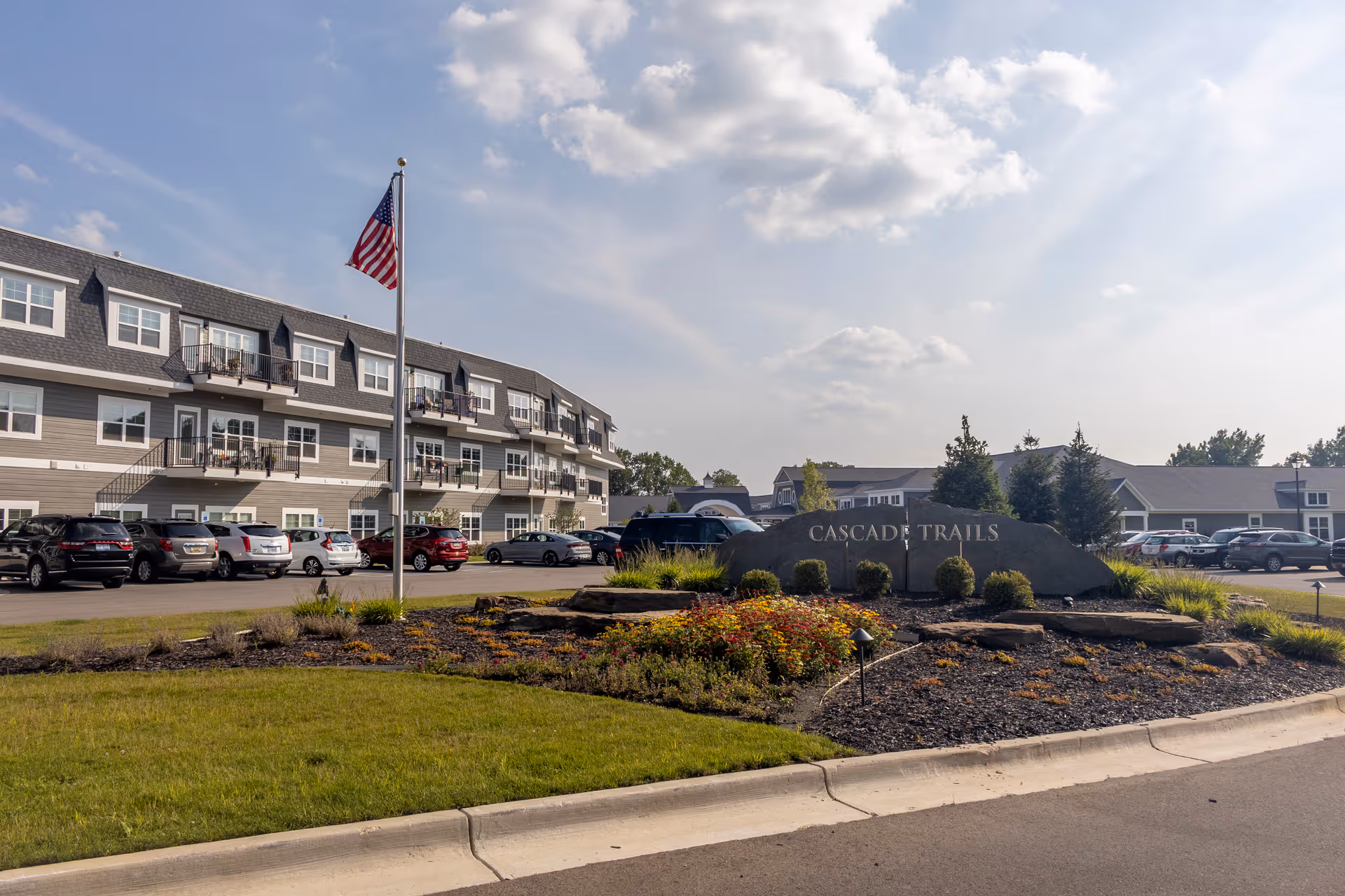 Exterior view of Cascade Trails Senior Living facility showing a multi-story building with balconies, a landscaped area with flowers and shrubs, an American flag on a flagpole, and a parking lot with several cars under a partly cloudy sky.