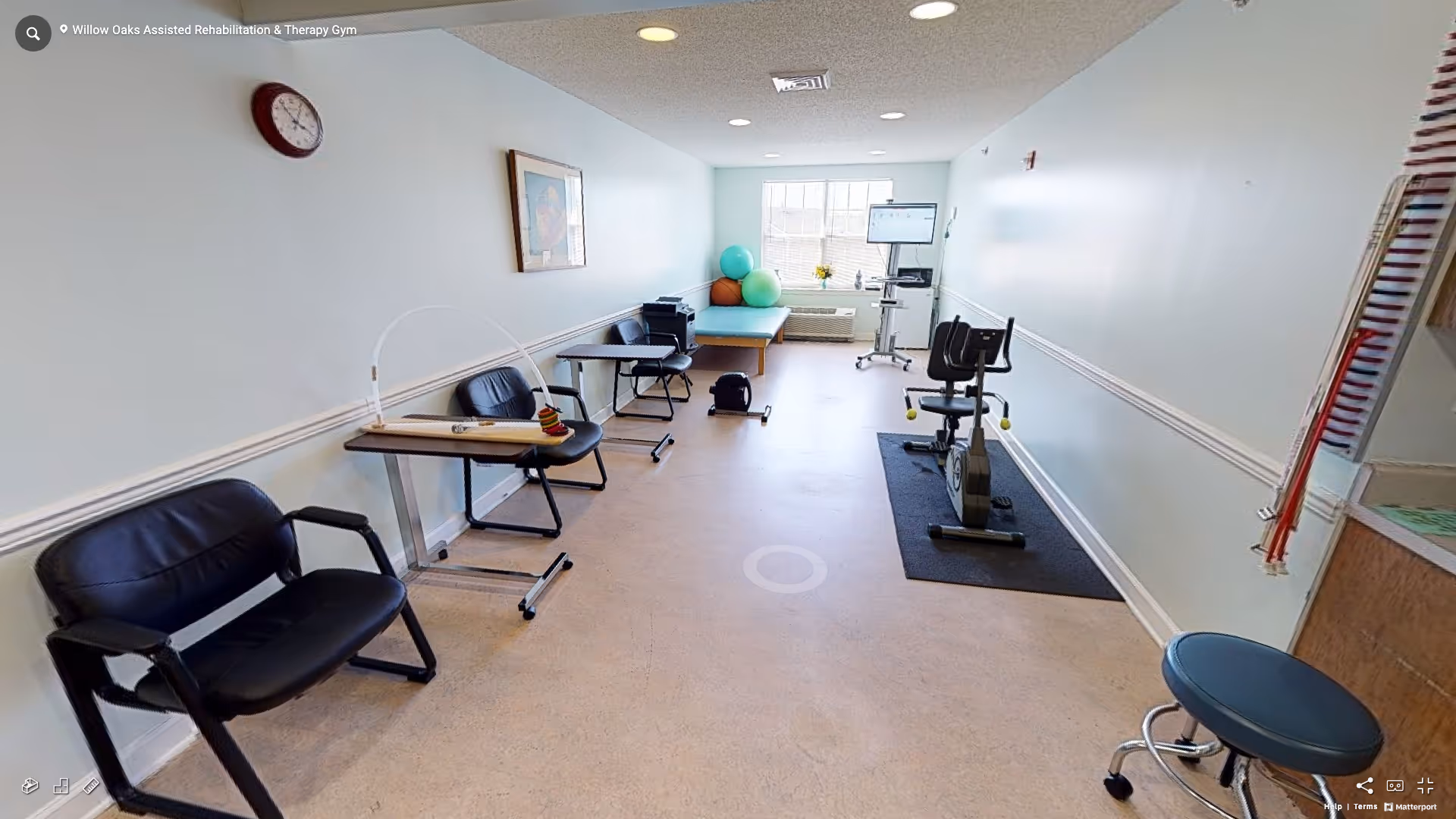 A rehabilitation and therapy gym room with exercise equipment including a stationary bike, therapy tables, chairs, exercise balls near a window, and a wall clock. The room has light-colored walls and flooring, with a framed picture on the wall and a stool near the entrance.