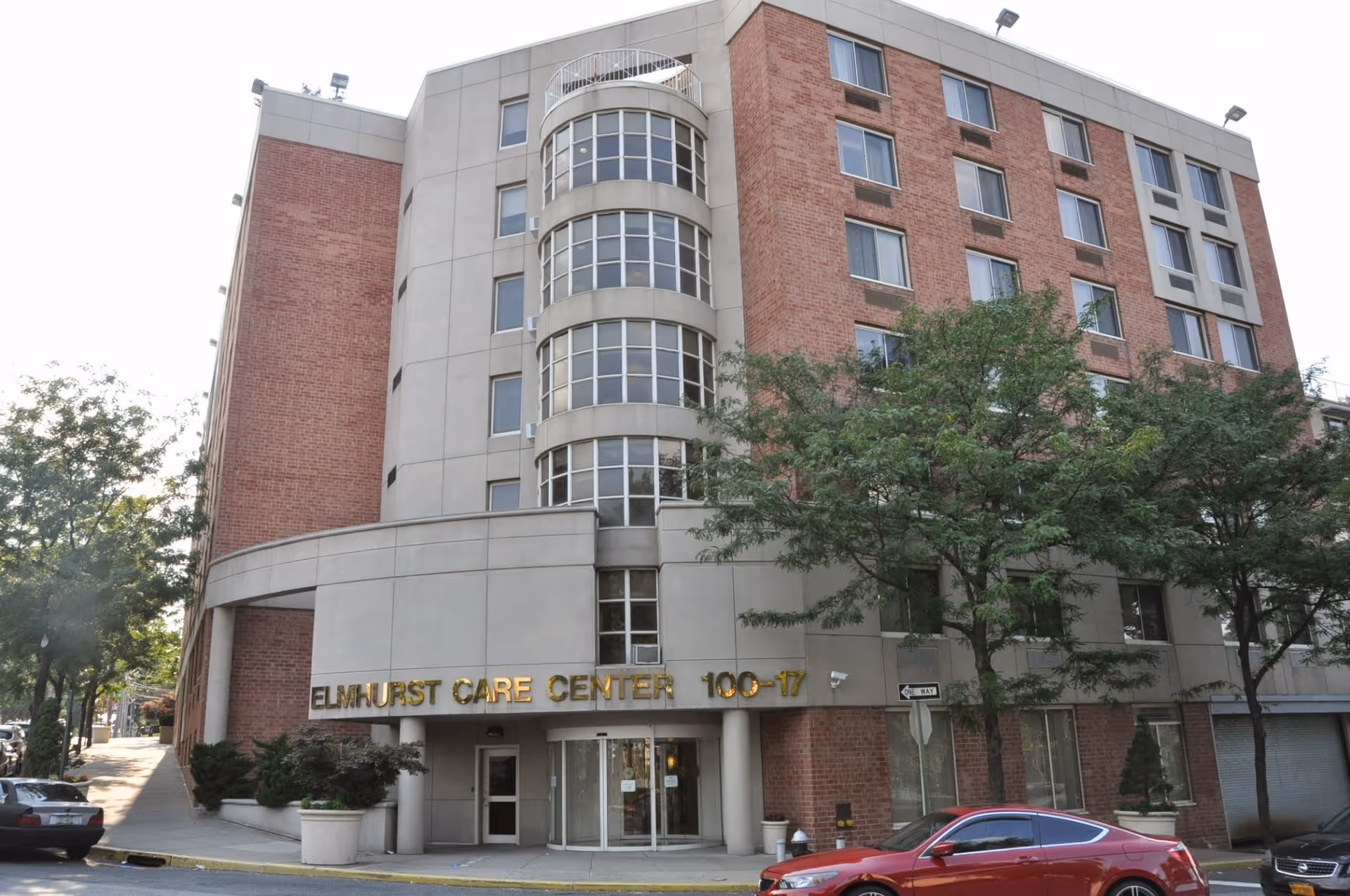 Exterior view of Elmhurst Care Center, a multi-story building with a combination of brick and concrete facade. The entrance is marked with gold lettering displaying the facility name and address number 100-17. Trees and parked cars are visible in front of the building.