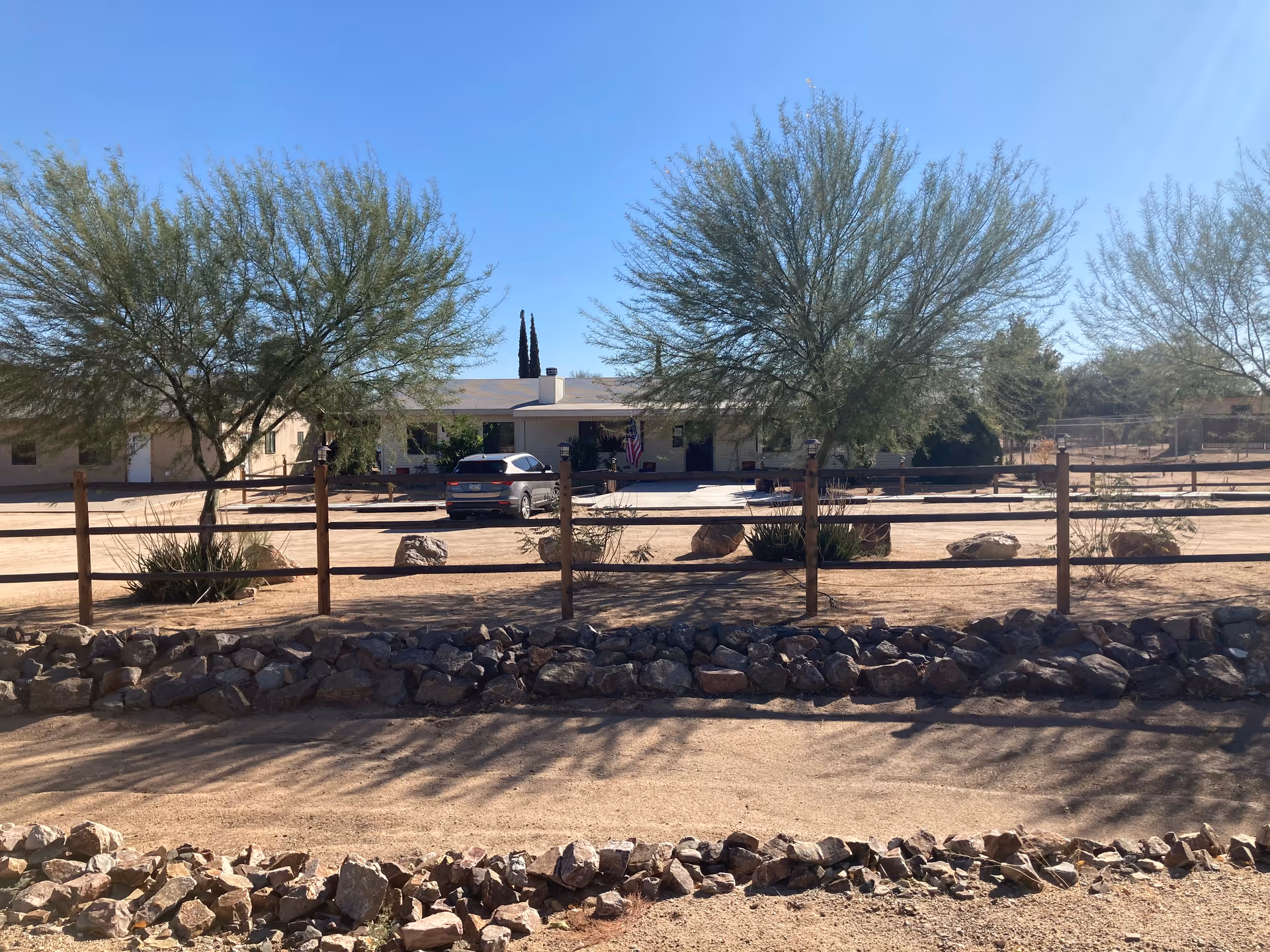 Single-story assisted living building behind a wooden fence with desert landscaping, trees, and a parked car under a clear blue sky.