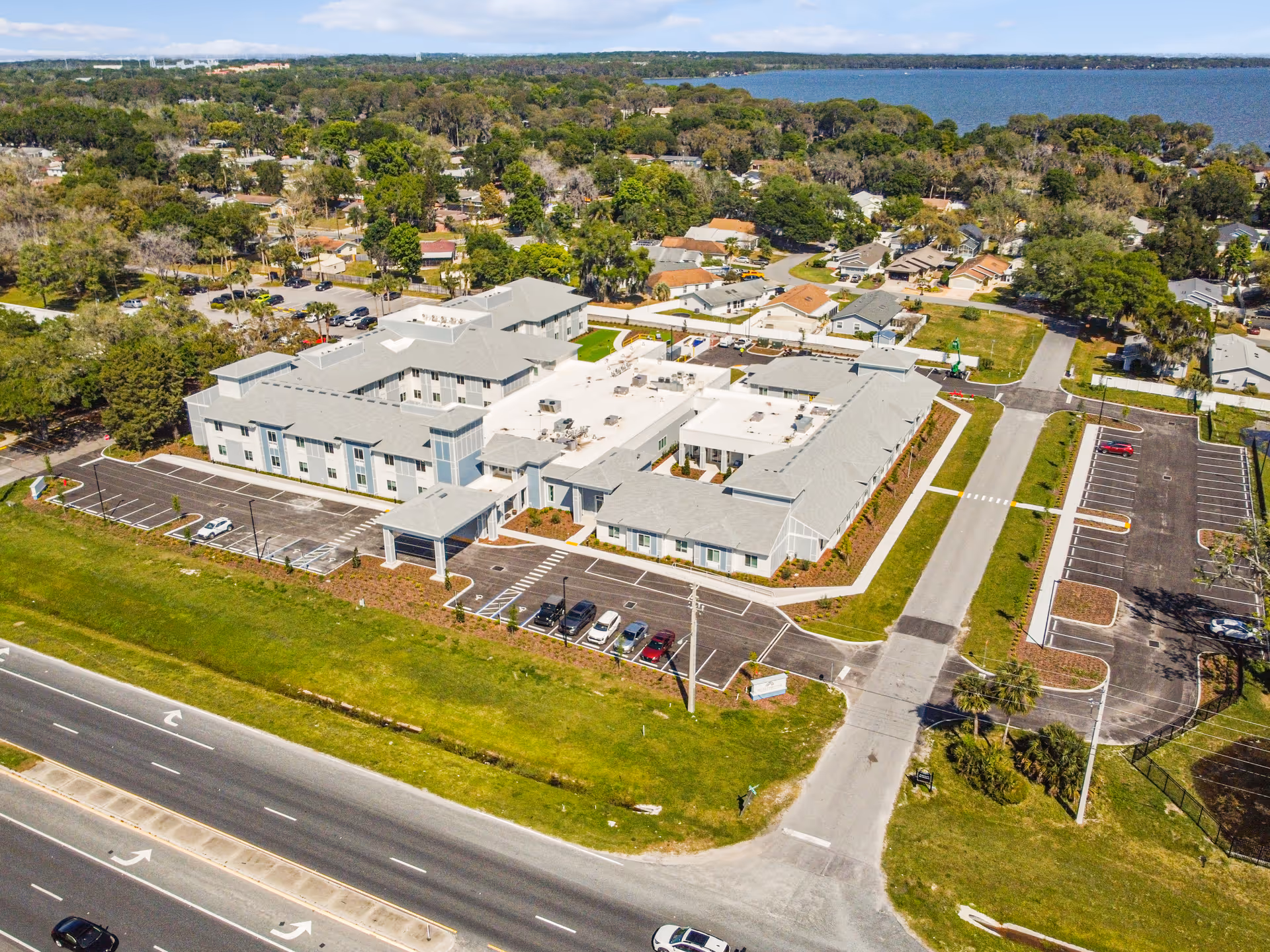 Aerial view of a large light-gray building complex with parking lots, entrance drive, a nearby roadway, and a lake in the background.