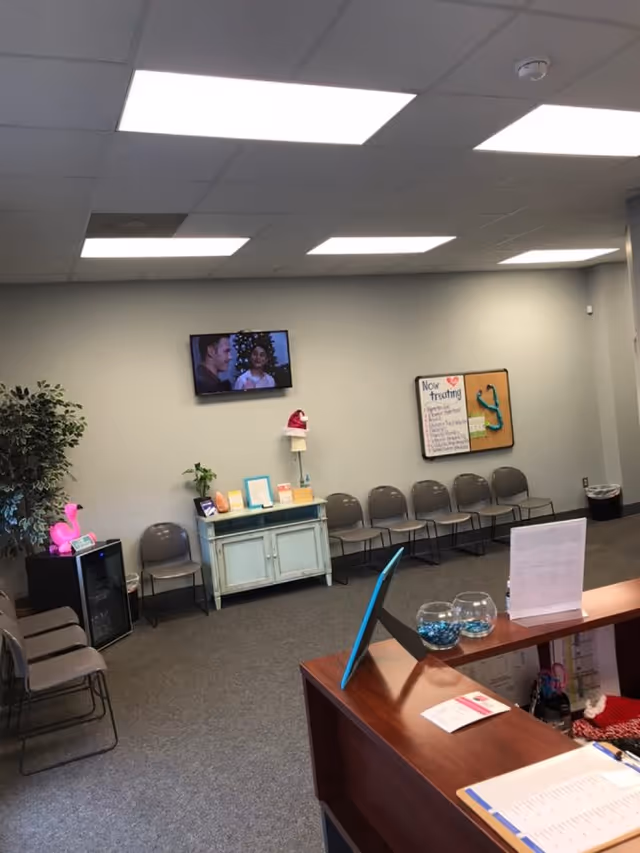 Reception area with rows of chairs, front desk, TV on wall, bulletin board.
