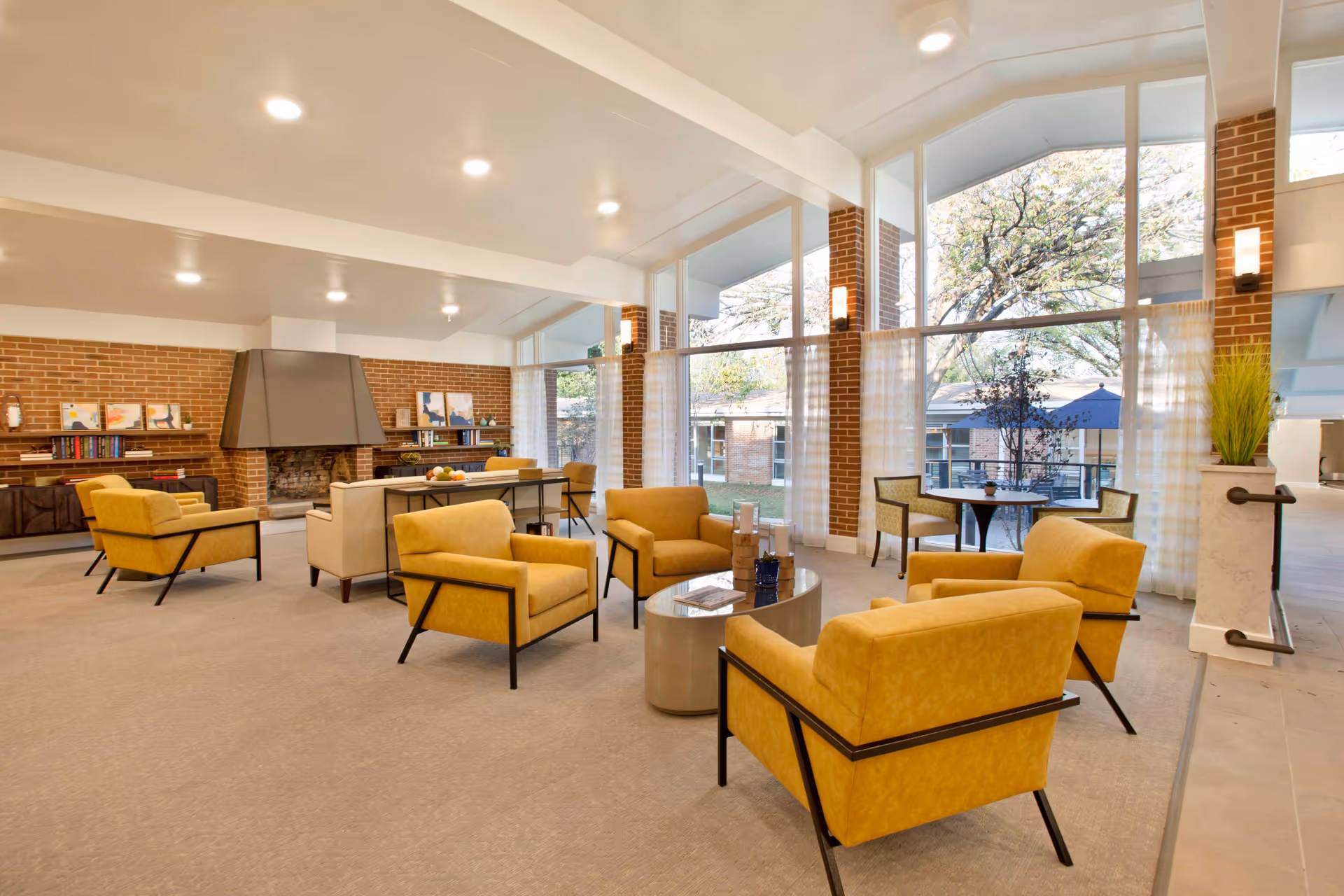 Bright senior living lounge with yellow armchairs, tables, fireplace, and large front windows.