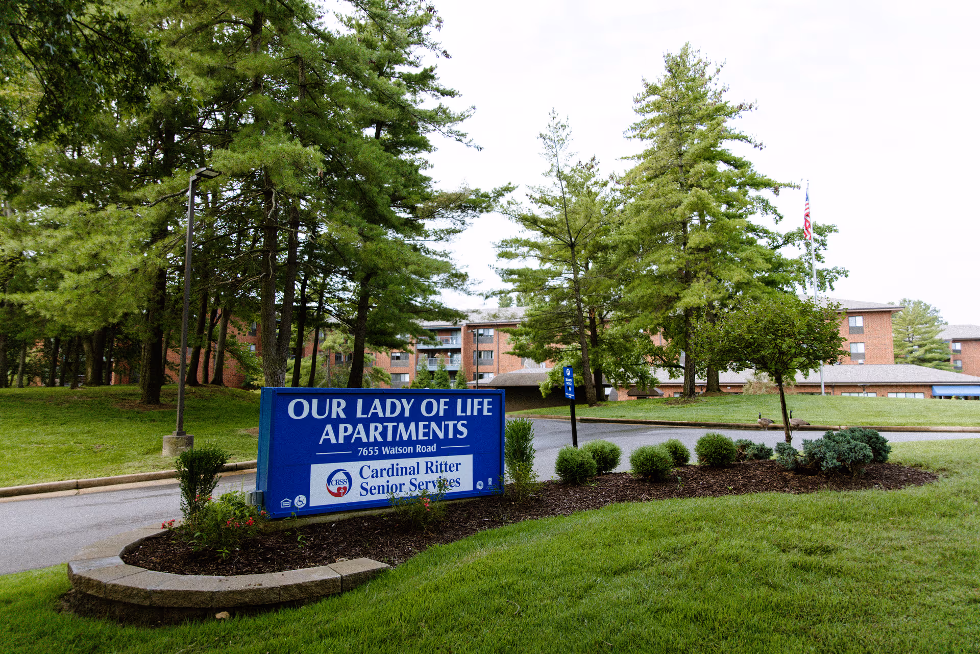 A blue sign for Our Lady of Life Apartments in front of a brick senior services building with green lawn and trees.