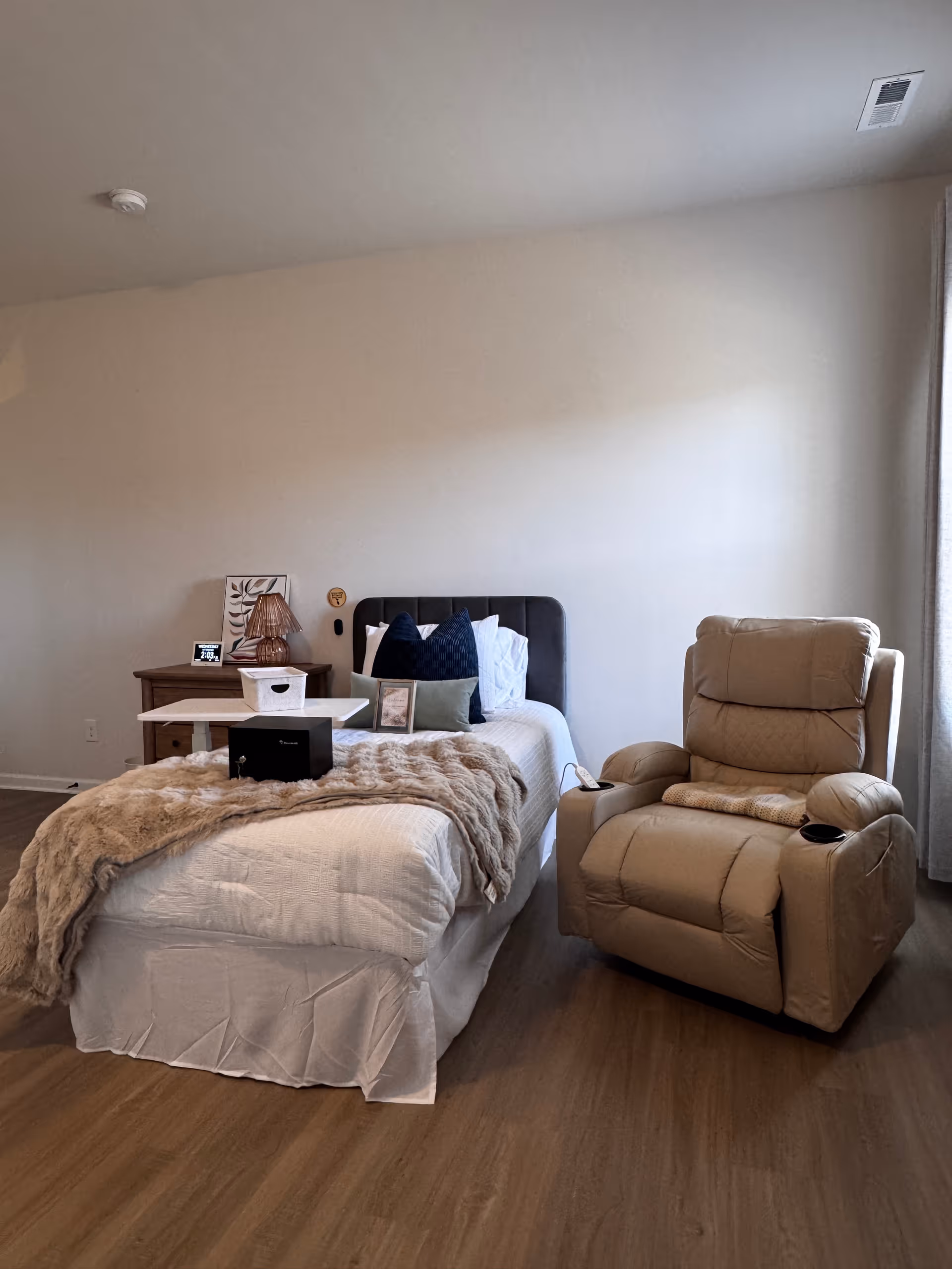 A cozy bedroom in a senior living facility with bed, beige recliner, nightstand.