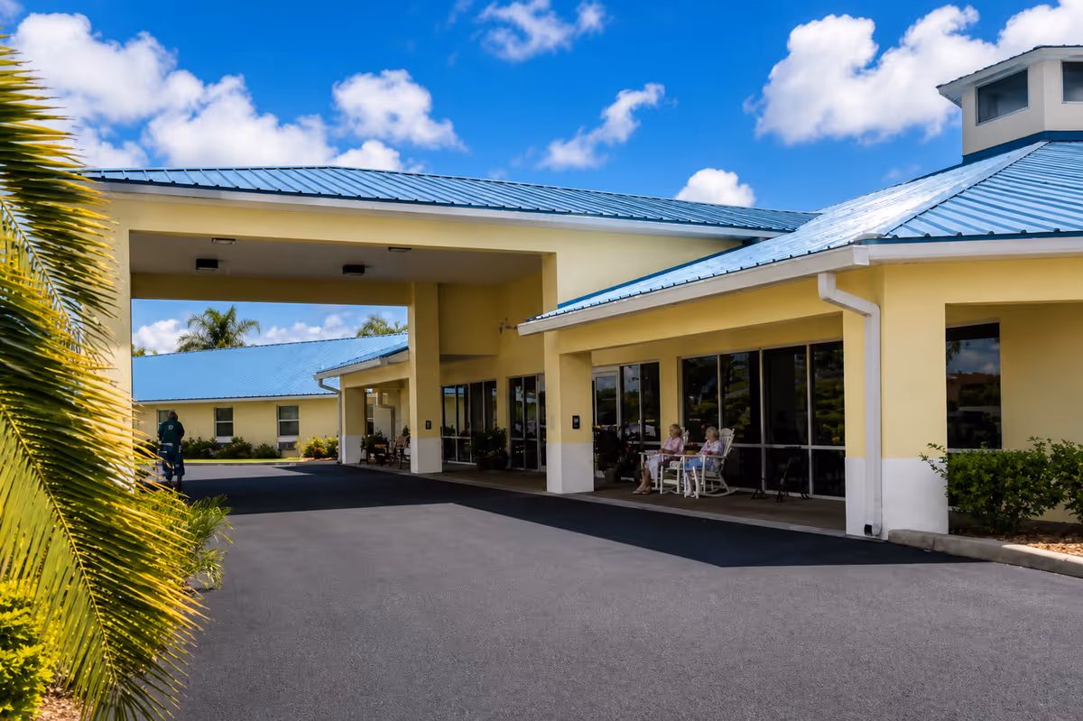 A yellow assisted living facility with a blue metal roof and covered entrance; residents seated outside.