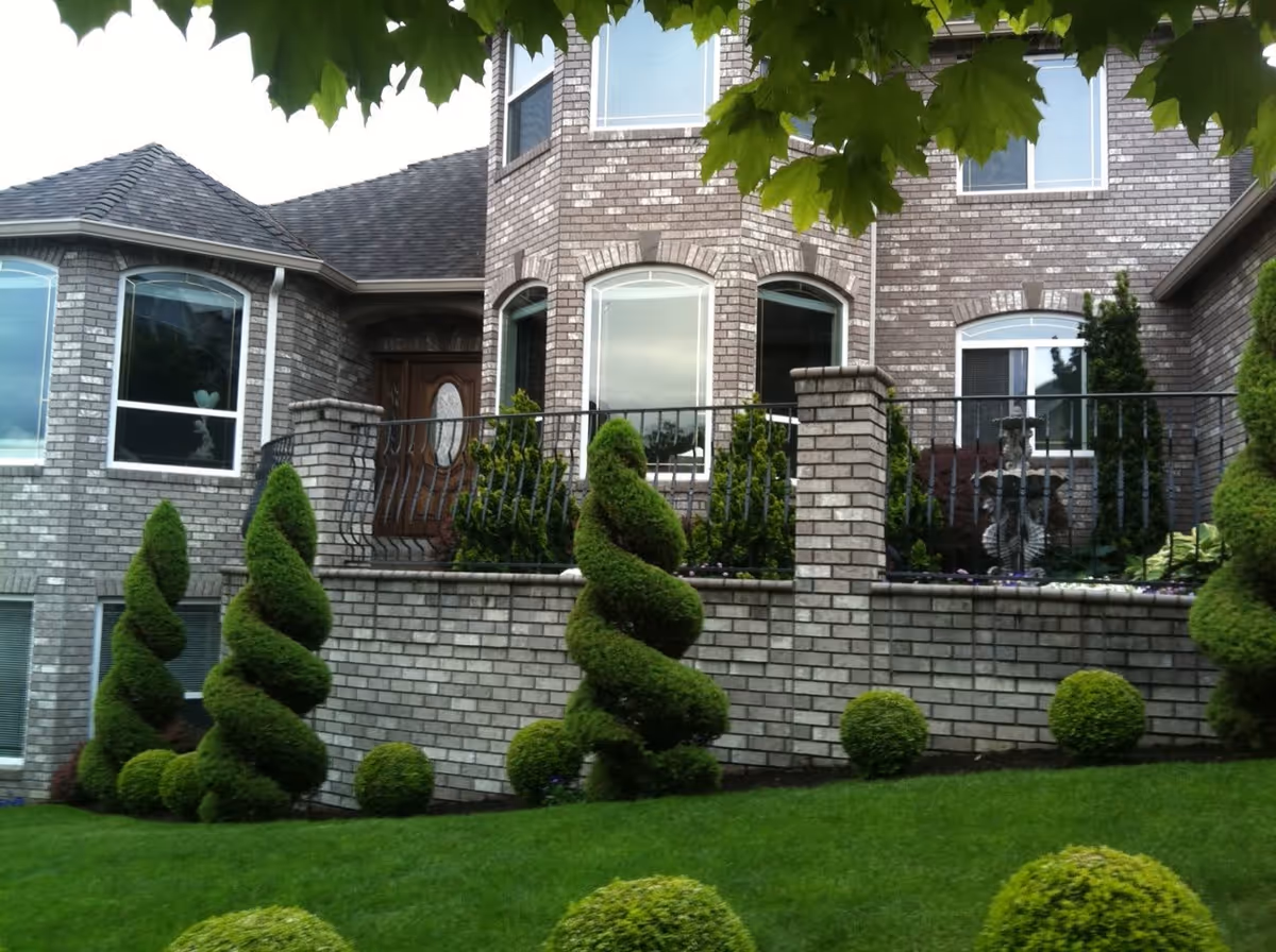 Front exterior view of a large brick building with multiple windows and a wooden front door. The building is surrounded by a well-maintained garden featuring neatly trimmed spiral and spherical bushes, a green lawn, and a black metal railing atop a brick retaining wall. Tree leaves partially frame the top of the image.