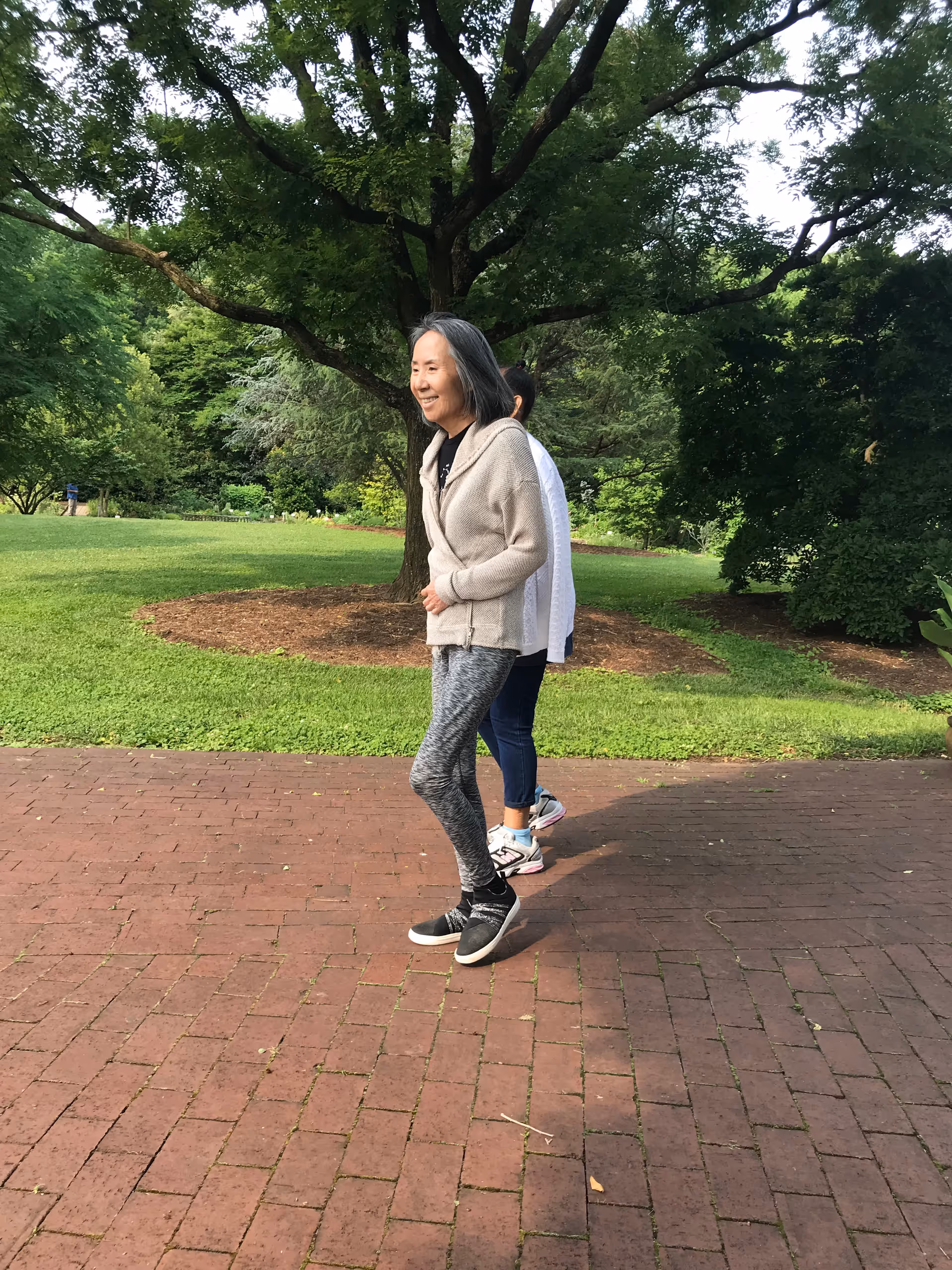 Two women walking on a brick pathway in a park-like outdoor setting with green grass and large trees in the background. One woman is smiling and wearing a light-colored jacket and patterned leggings, while the other is partially obscured behind her.