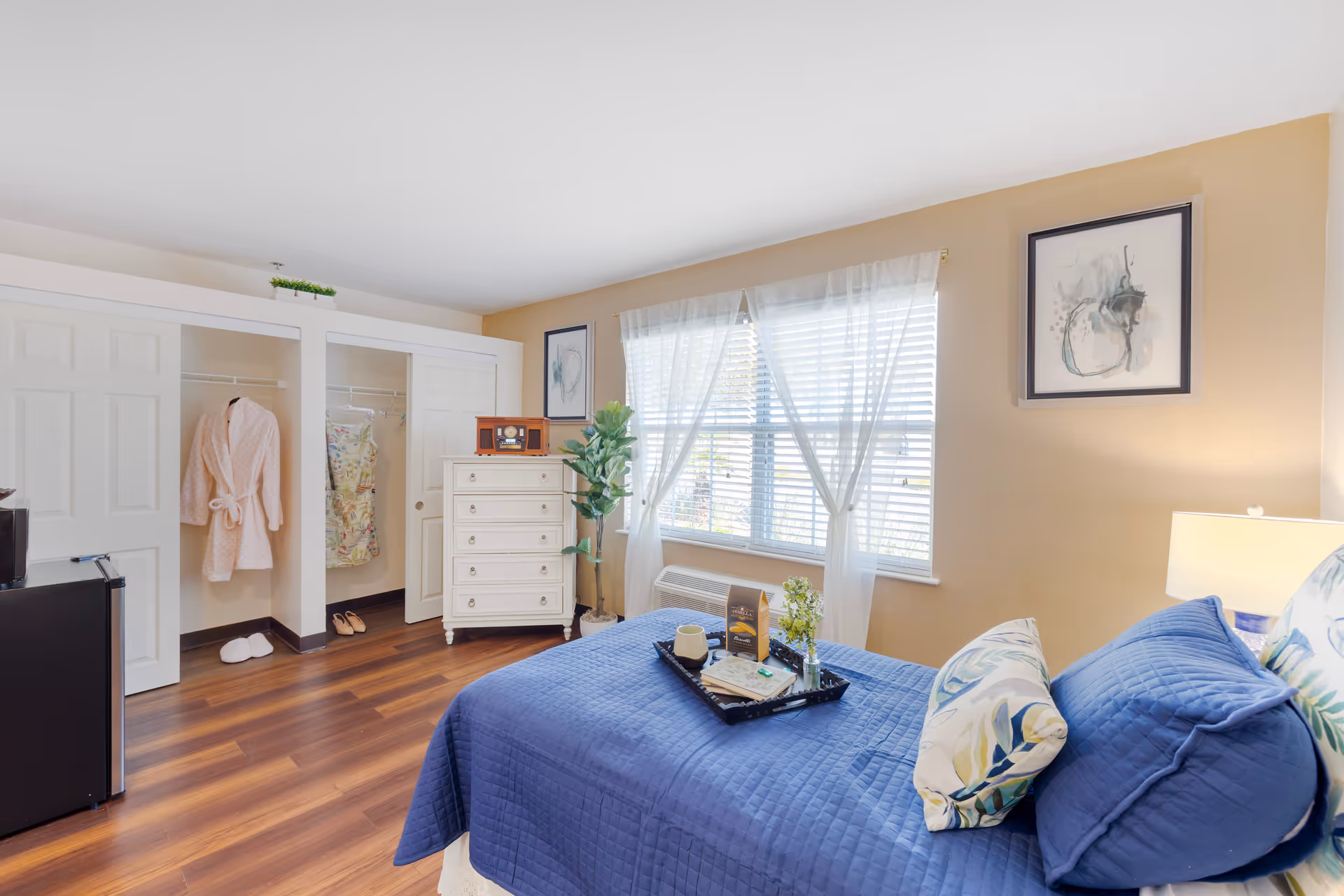 A bright bedroom with a bed covered in a blue quilt and floral pillows. A tray with a cup, book, and small plant is on the bed. There is a window with white sheer curtains letting in natural light. A white dresser with a vintage radio on top stands next to a green potted plant. Two open closets show hanging clothes, slippers, and a robe. The floor is wood, and the walls are painted beige with two framed abstract artworks.
