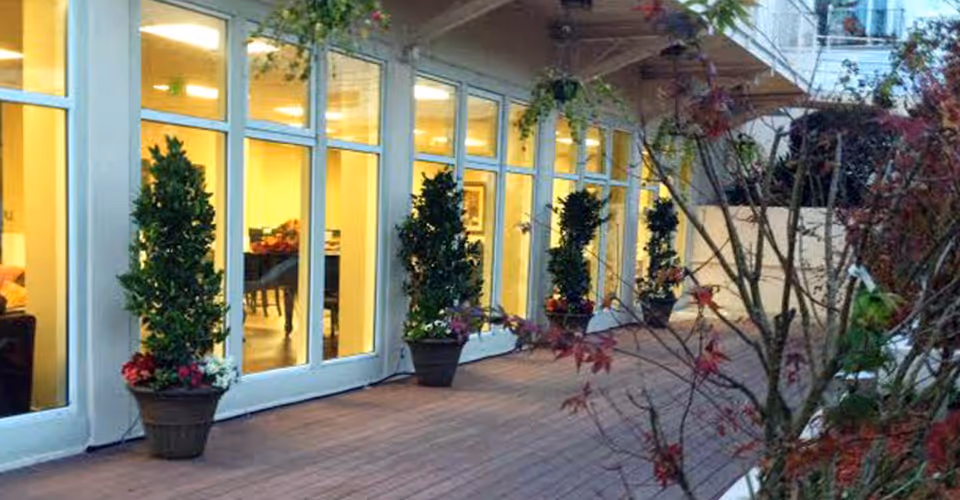 Outdoor patio with potted plants lined up against large glass windows showing warm interior lighting.