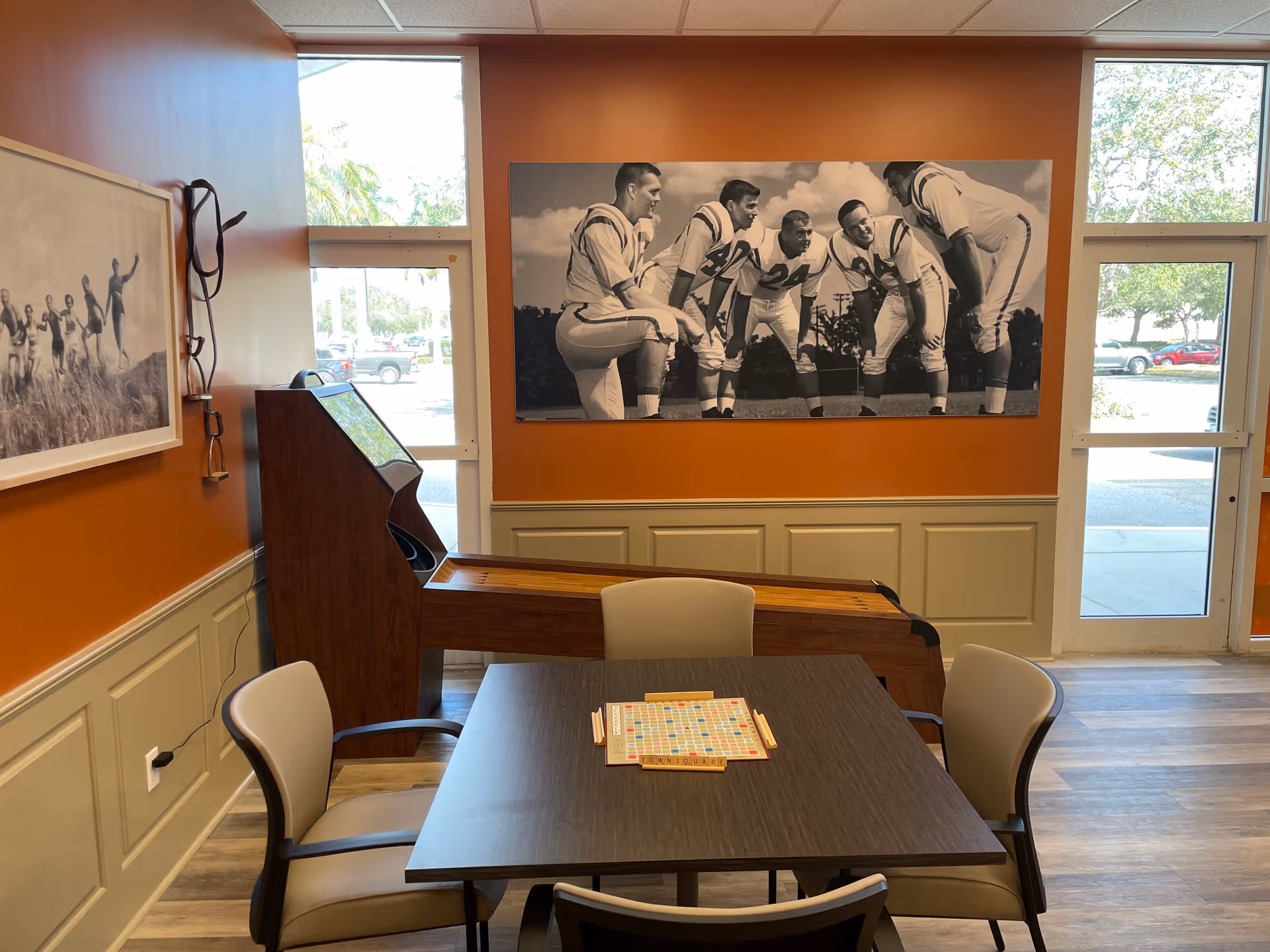 A small communal dining and activity area with a square table, chairs, and orange wall art.