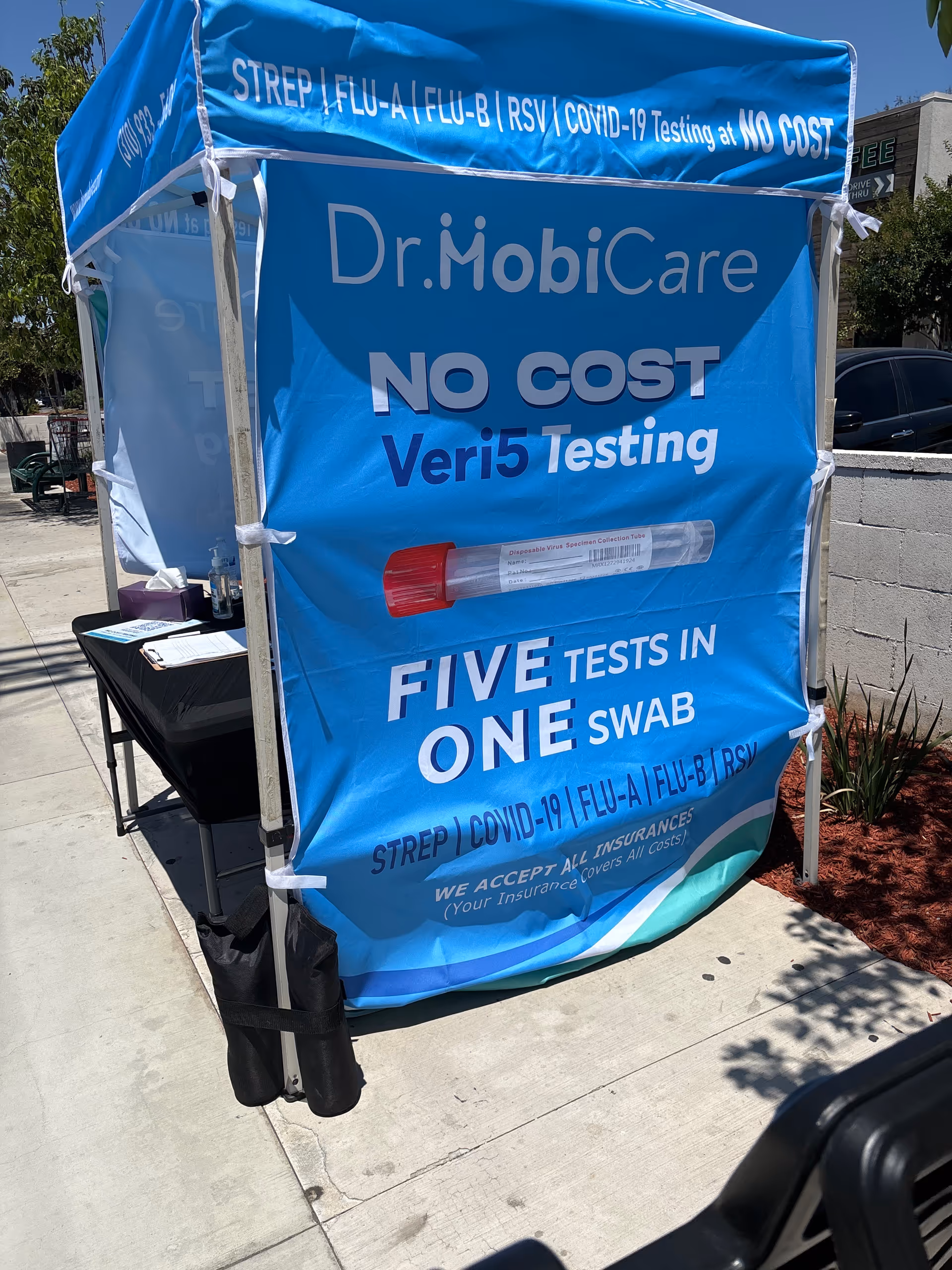 Blue Dr. MobiCare outdoor testing tent advertising no-cost COVID-19 tests with a table nearby.