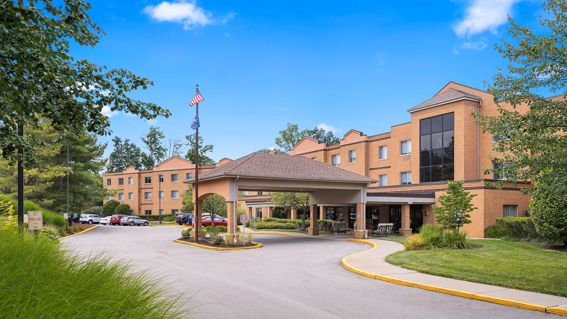 Exterior view of Holiday Niagara Village senior living facility showing a large brick building with multiple windows, a covered entrance driveway, parked cars, green lawns, trees, and a clear blue sky with some clouds.