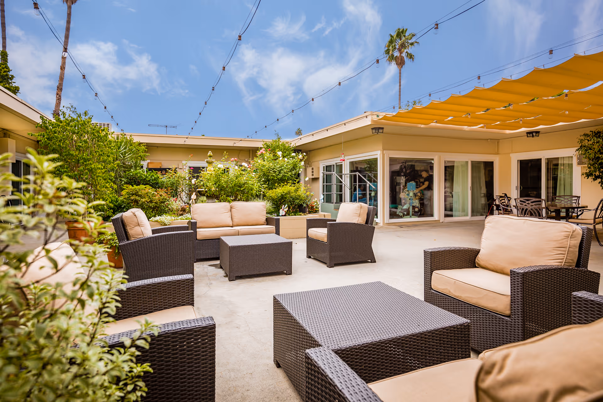 Outdoor patio area at Brentwood Health Care Center featuring cushioned wicker chairs and sofas arranged around matching coffee tables. The space is decorated with string lights overhead, yellow shade sails, and surrounded by greenery and potted plants. The building with large windows and sliding doors encloses the patio.